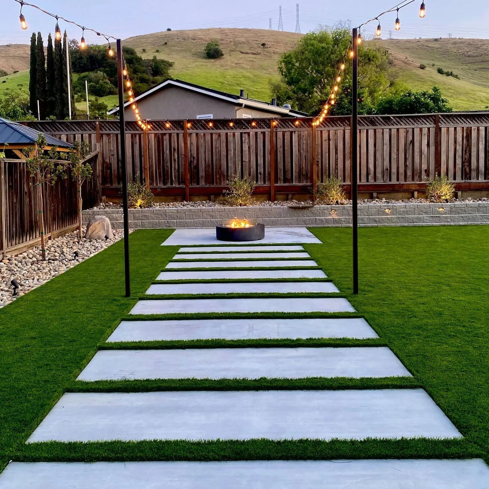 Backyard with a concrete walkway, fire pit, string lights, green lawn, and wooden fence.