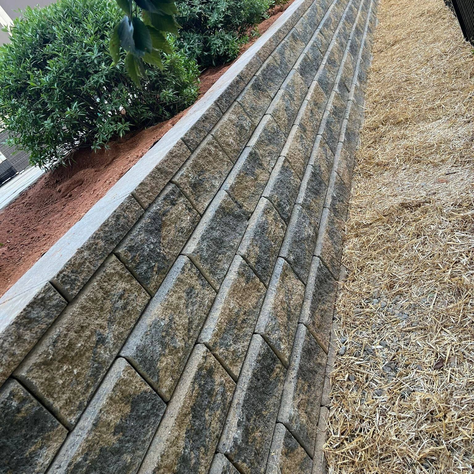 Retaining wall made of textured stone blocks, holding back a sloped landscape with mulch and greenery.