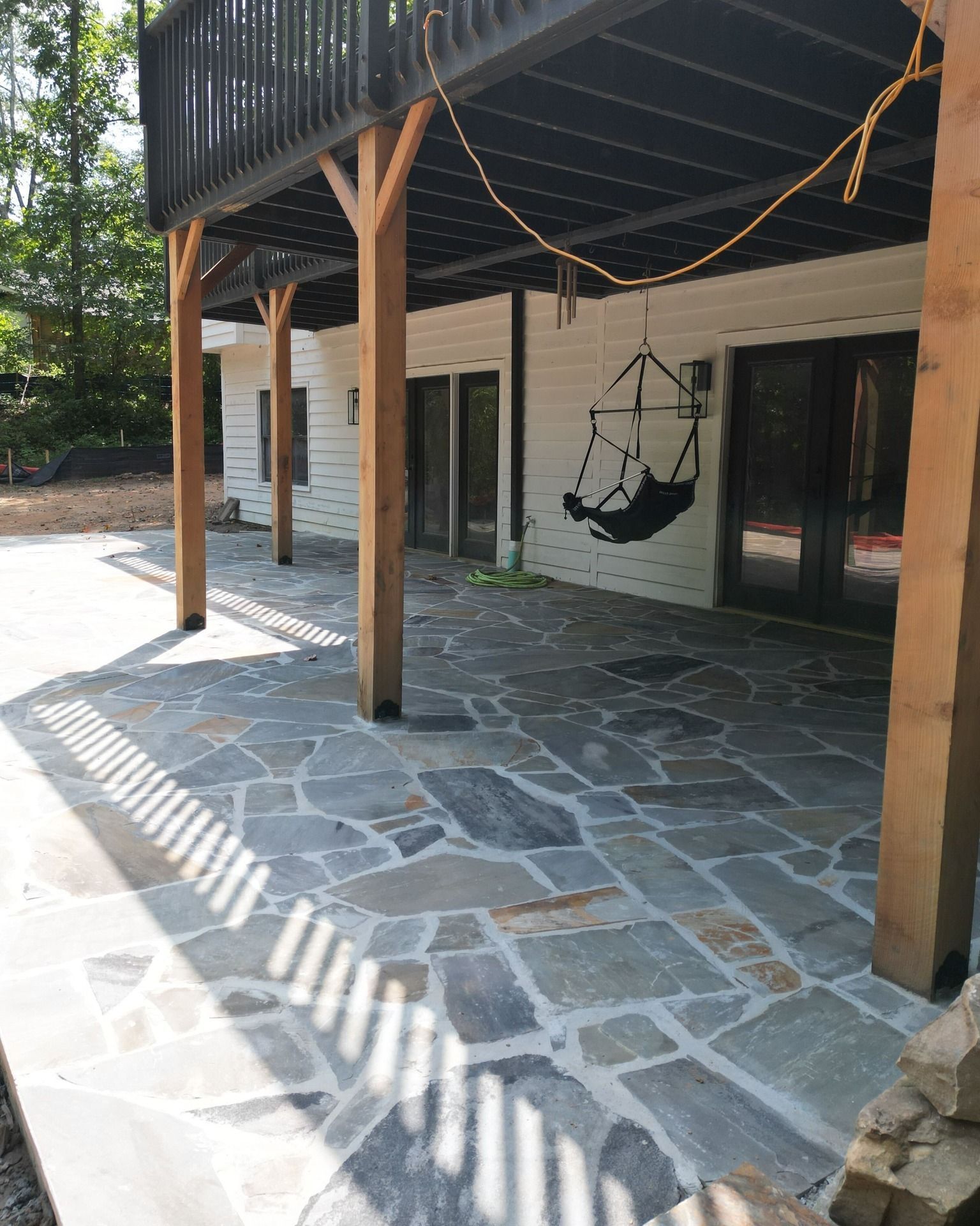 Flagstone patio under a deck with a hanging chair, wooden posts, and white house with glass doors.