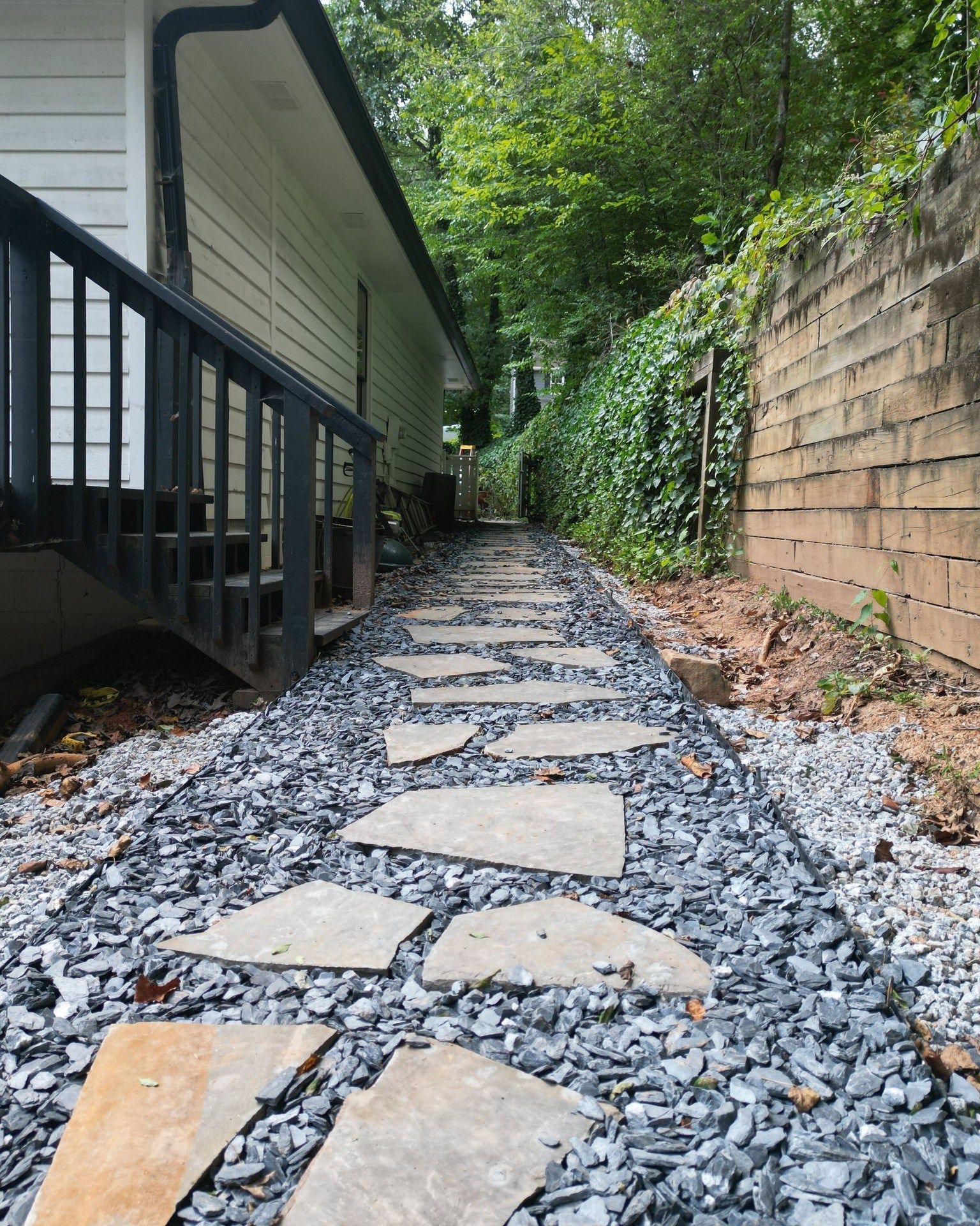 Stone path through a backyard, flanked by a house and ivy-covered brick wall. Gravel surrounds the stones.