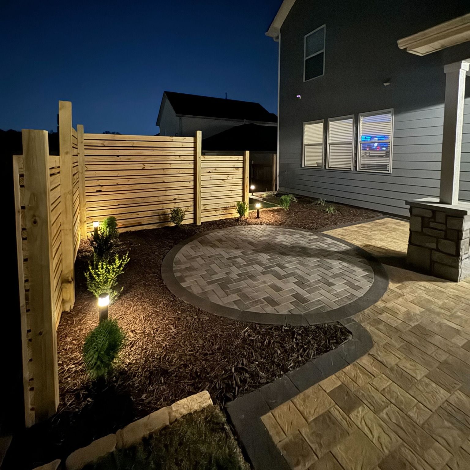 Backyard patio at night with lit pavers, a wood fence, and house.