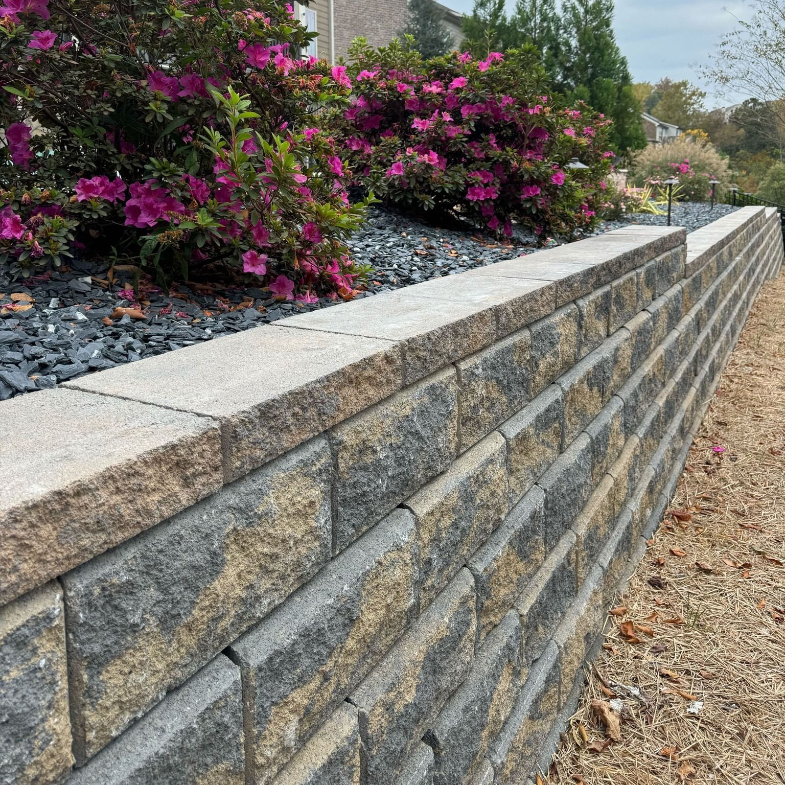 Stone retaining wall with blooming pink flowers on top, in an outdoor setting.