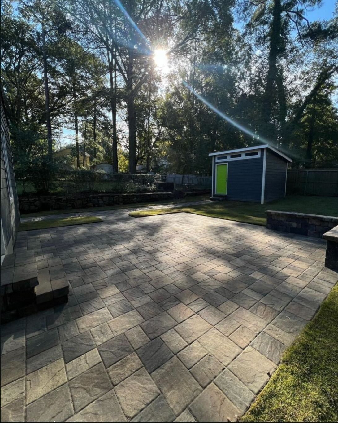 Brick patio in a backyard with a dark blue shed, green door, and trees in the background.