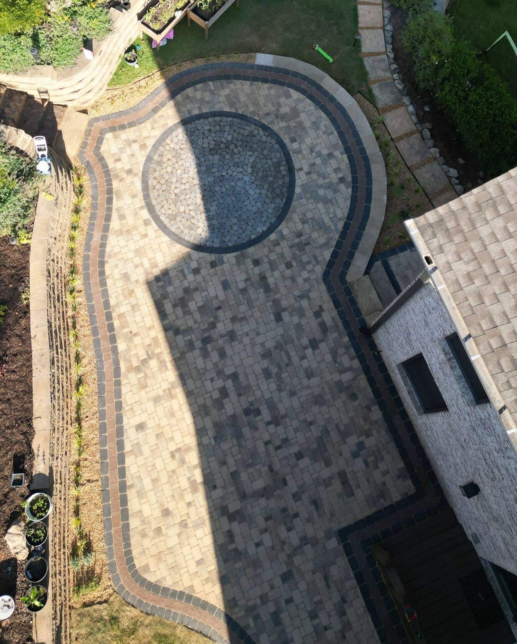 Overhead view of a patterned patio with a circular centerpiece, tan and dark bricks, next to a house and garden.