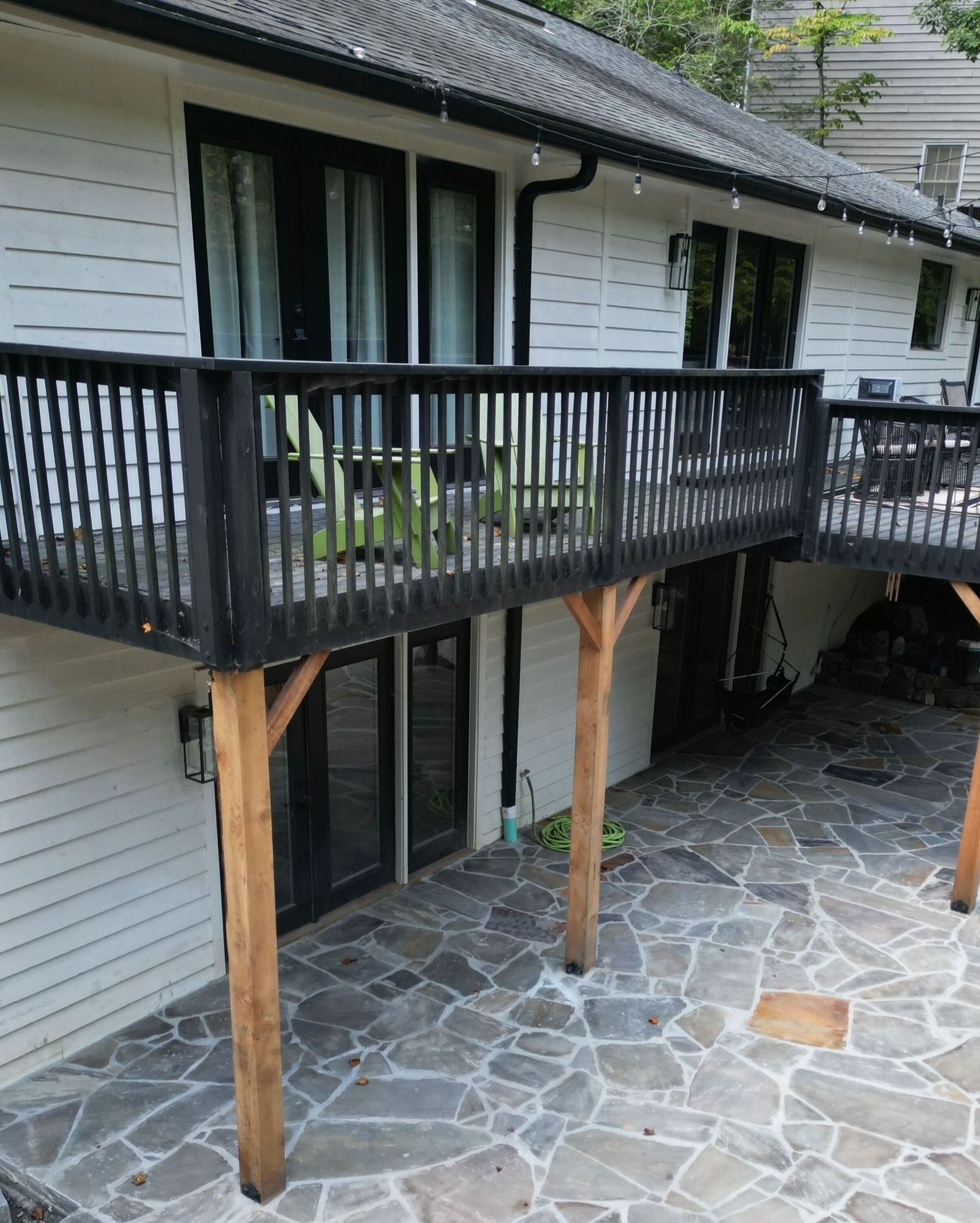 Black-railed deck supported by wooden posts, overlooking a flagstone patio. House is white with black trim.