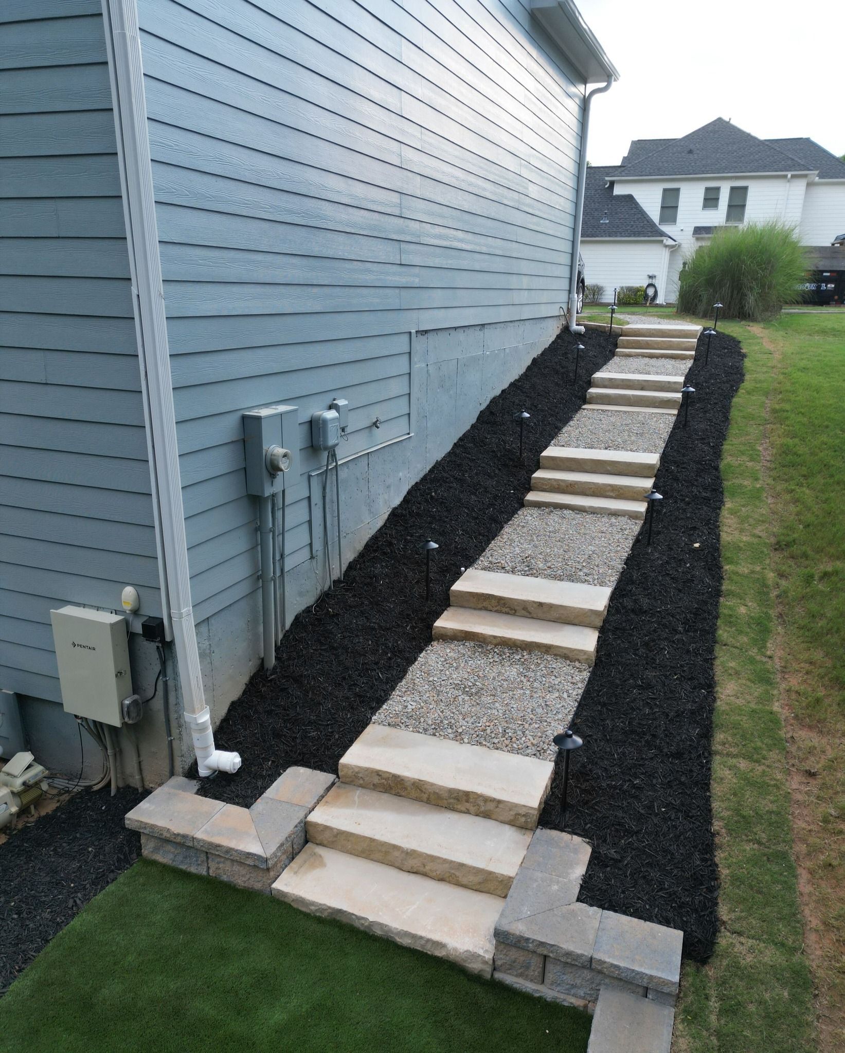 Stone steps with gravel pathway bordered by black mulch next to a blue house and green grass.