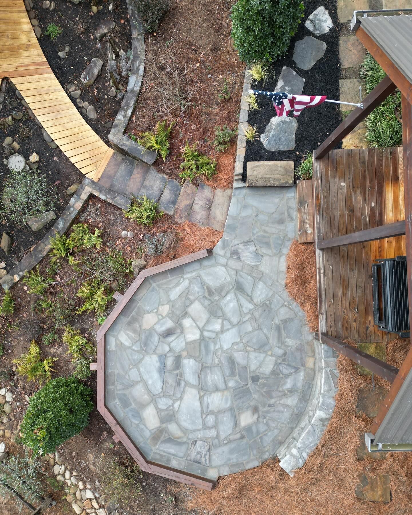 Overhead view of a stone patio, wooden walkway, stairs, and flag near a house with surrounding landscaping.
