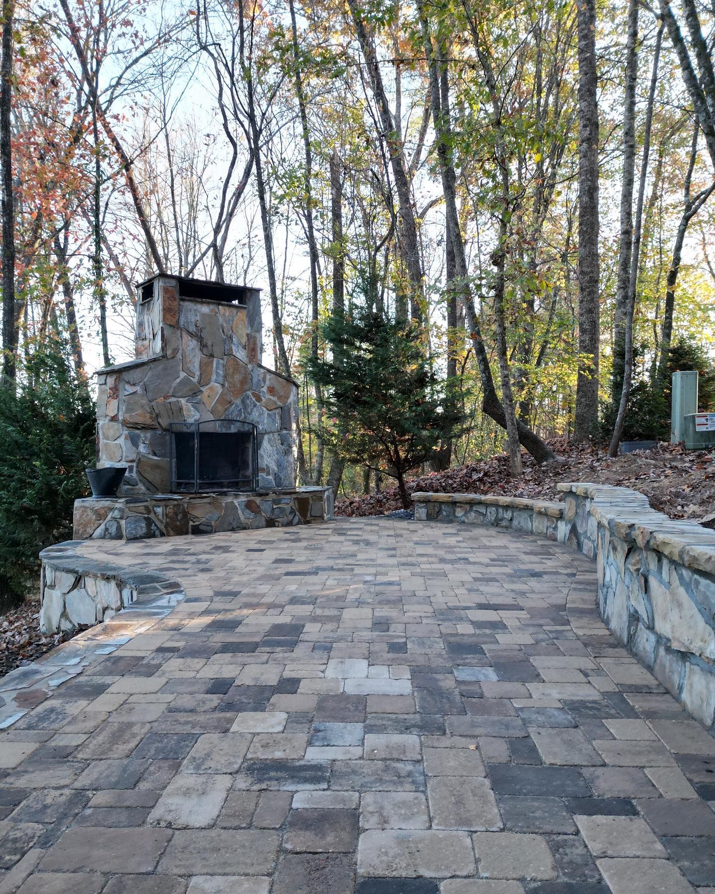 Stone patio with a fireplace in a wooded area.