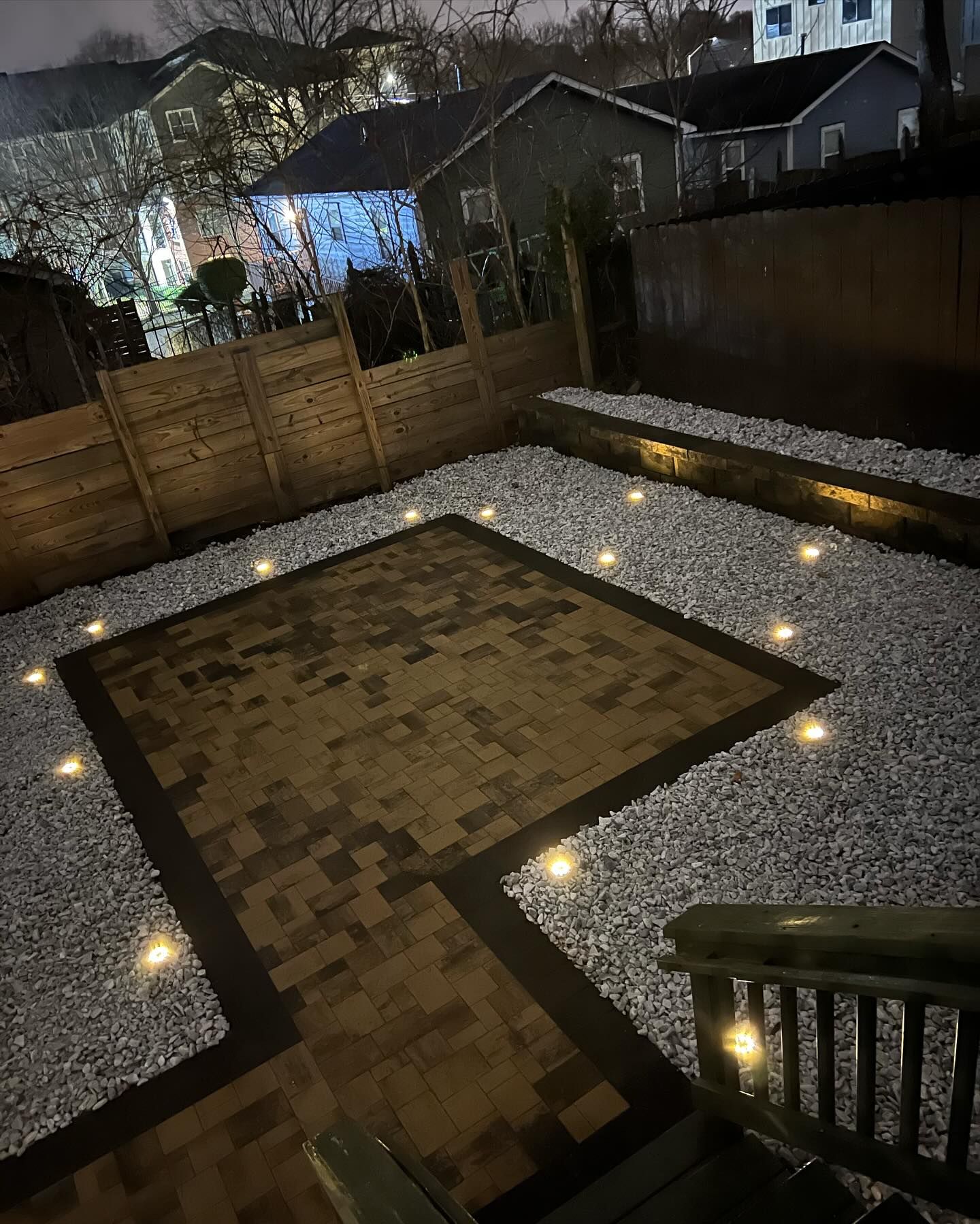 Backyard patio at night, brick pavers, white gravel, wooden fence, and small lights.