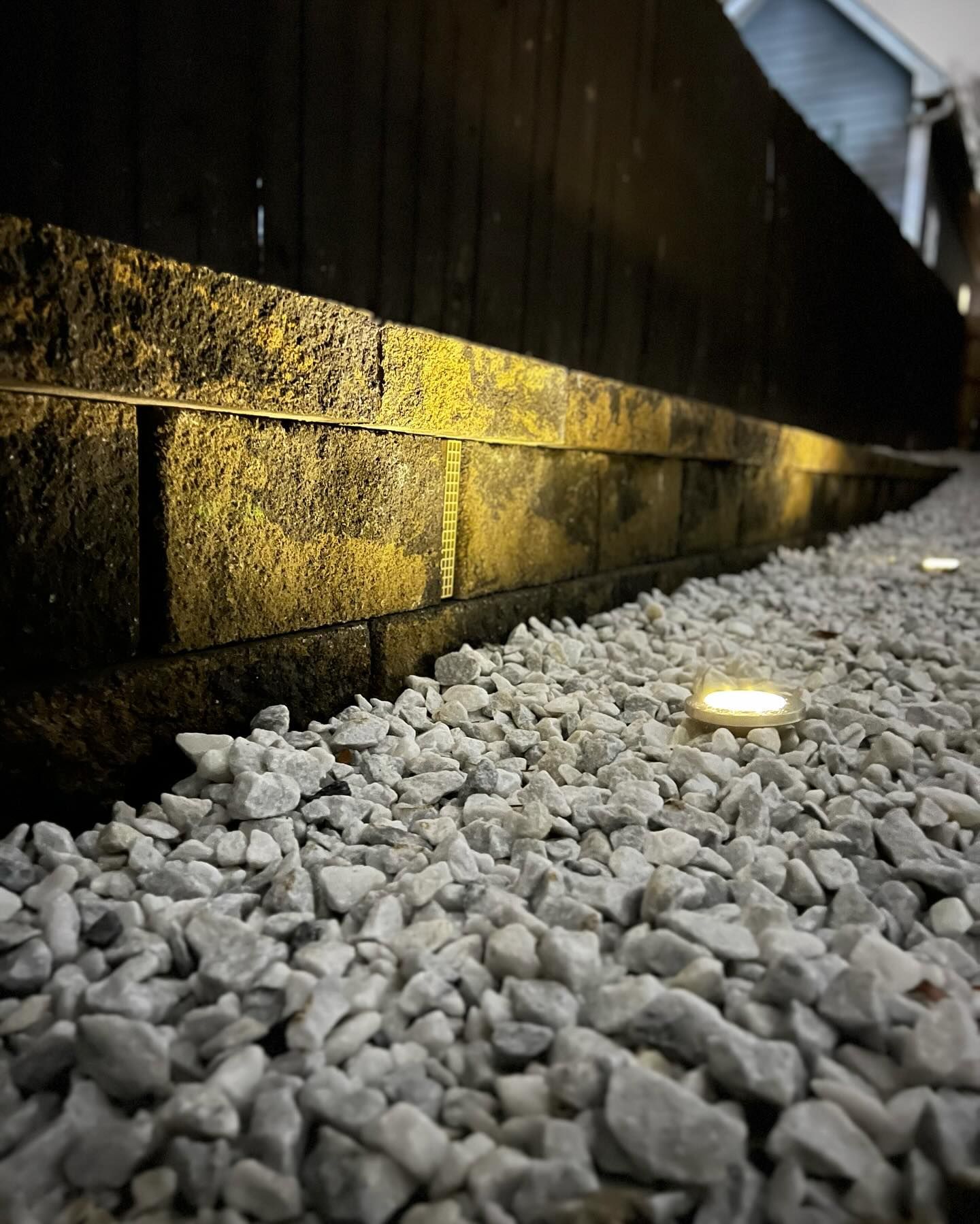 Gravel pathway illuminated by spotlights along a dark brick wall and wooden fence.