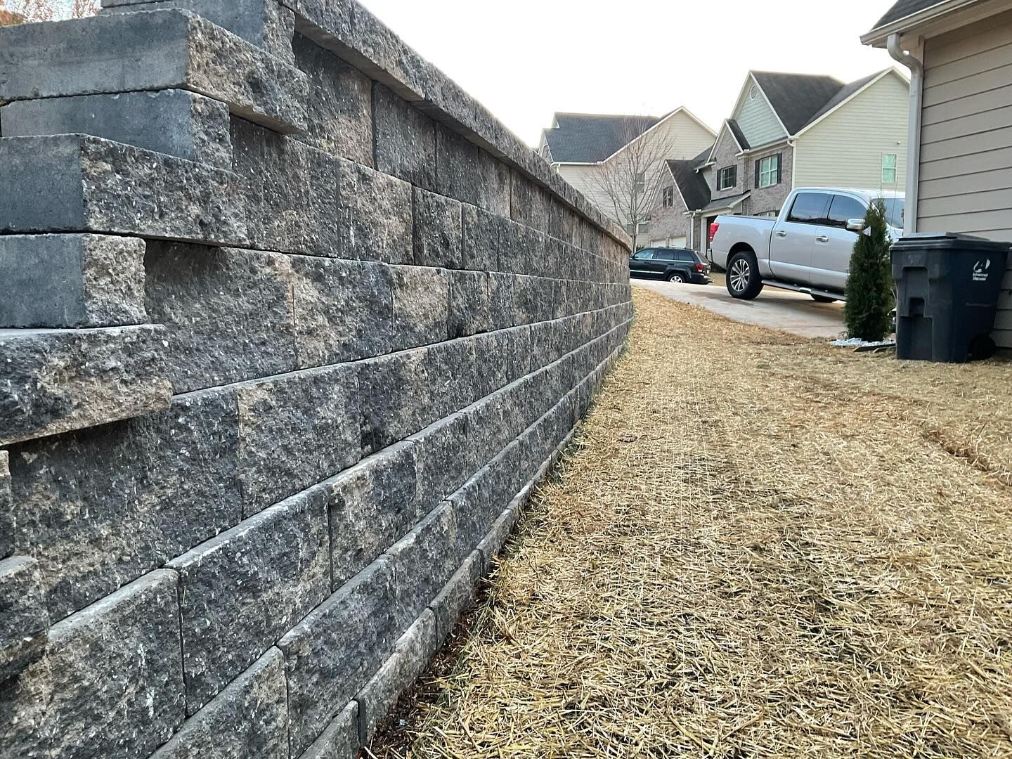 Retaining wall built with gray blocks beside a gravel area, houses and a truck in the background.