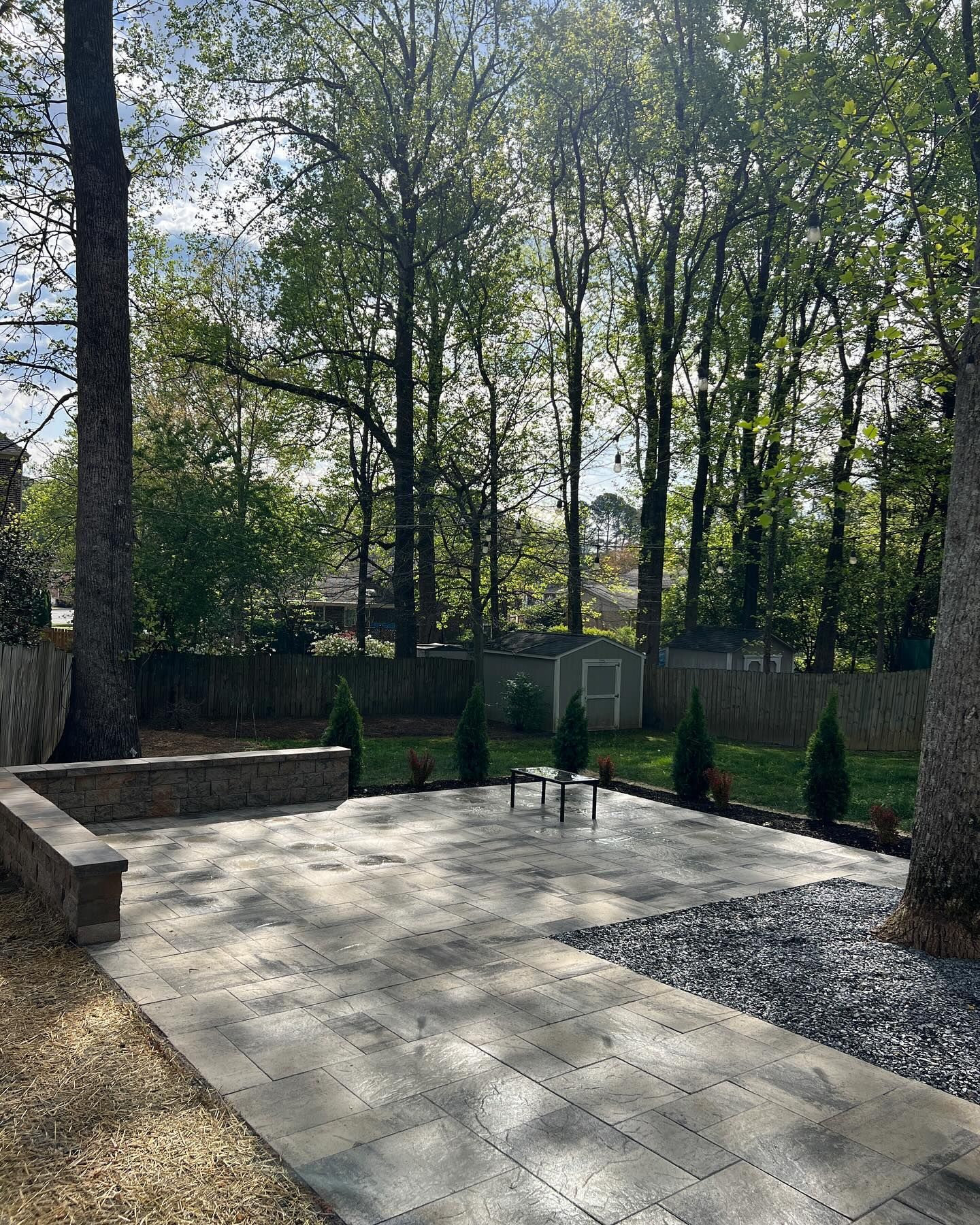 Patio with gray pavers, small evergreens, and trees. A small shed is visible in the background.