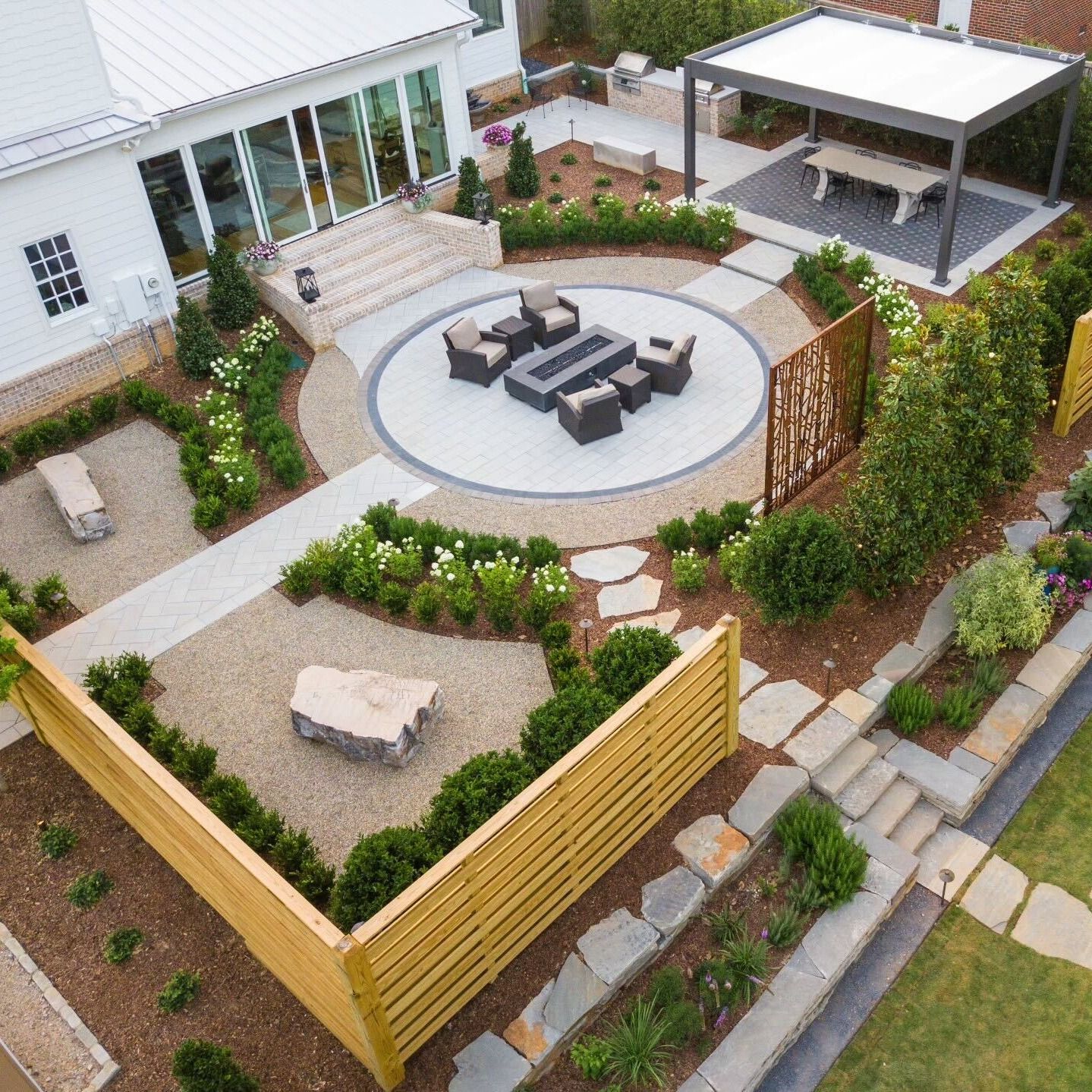 An aerial view of a modern backyard with a fire pit, dining pergola, stone paths, wooden fencing, and landscaped garden.