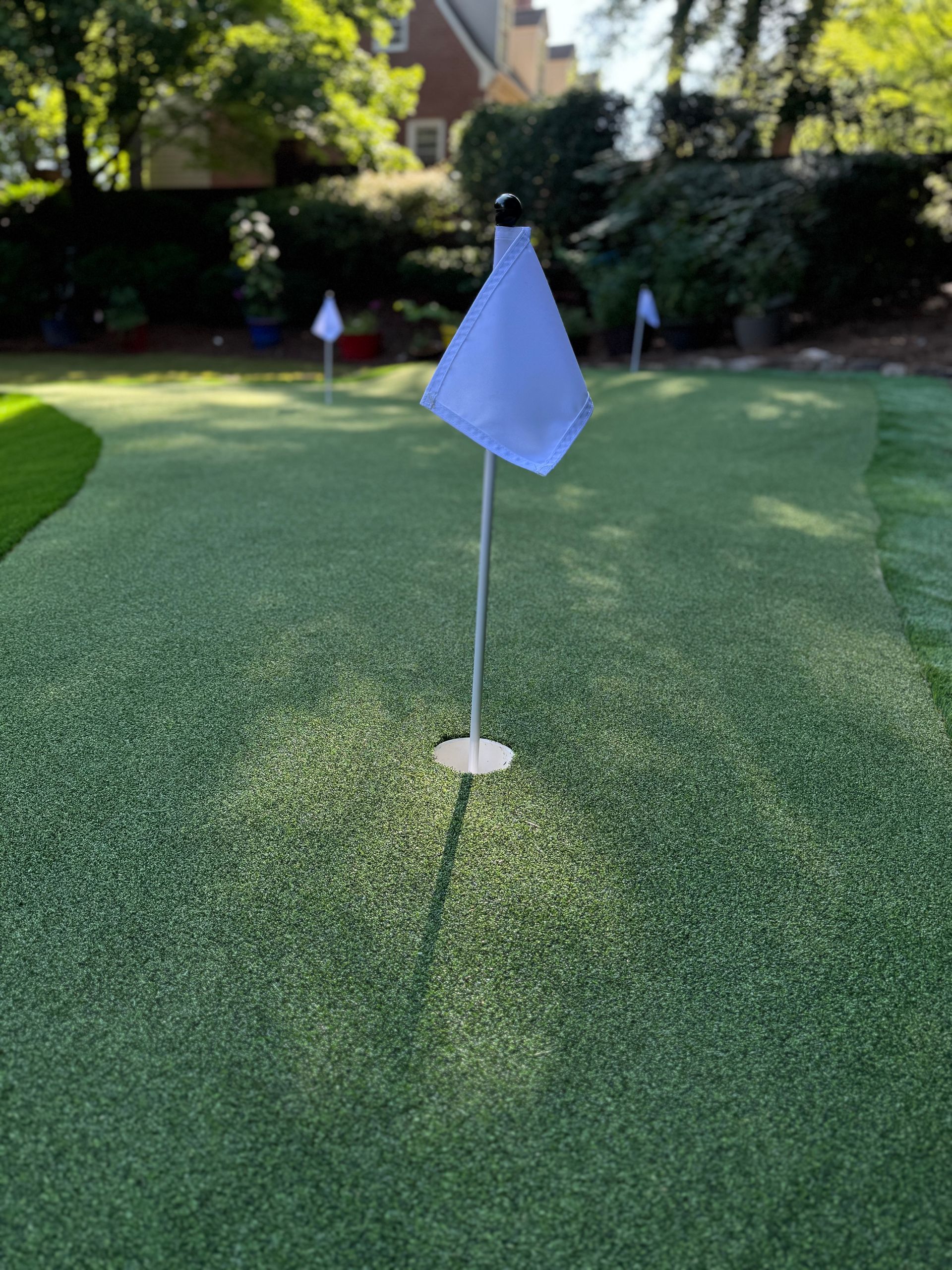 A green artificial putting green with a flagstick and white flag; in a backyard setting.