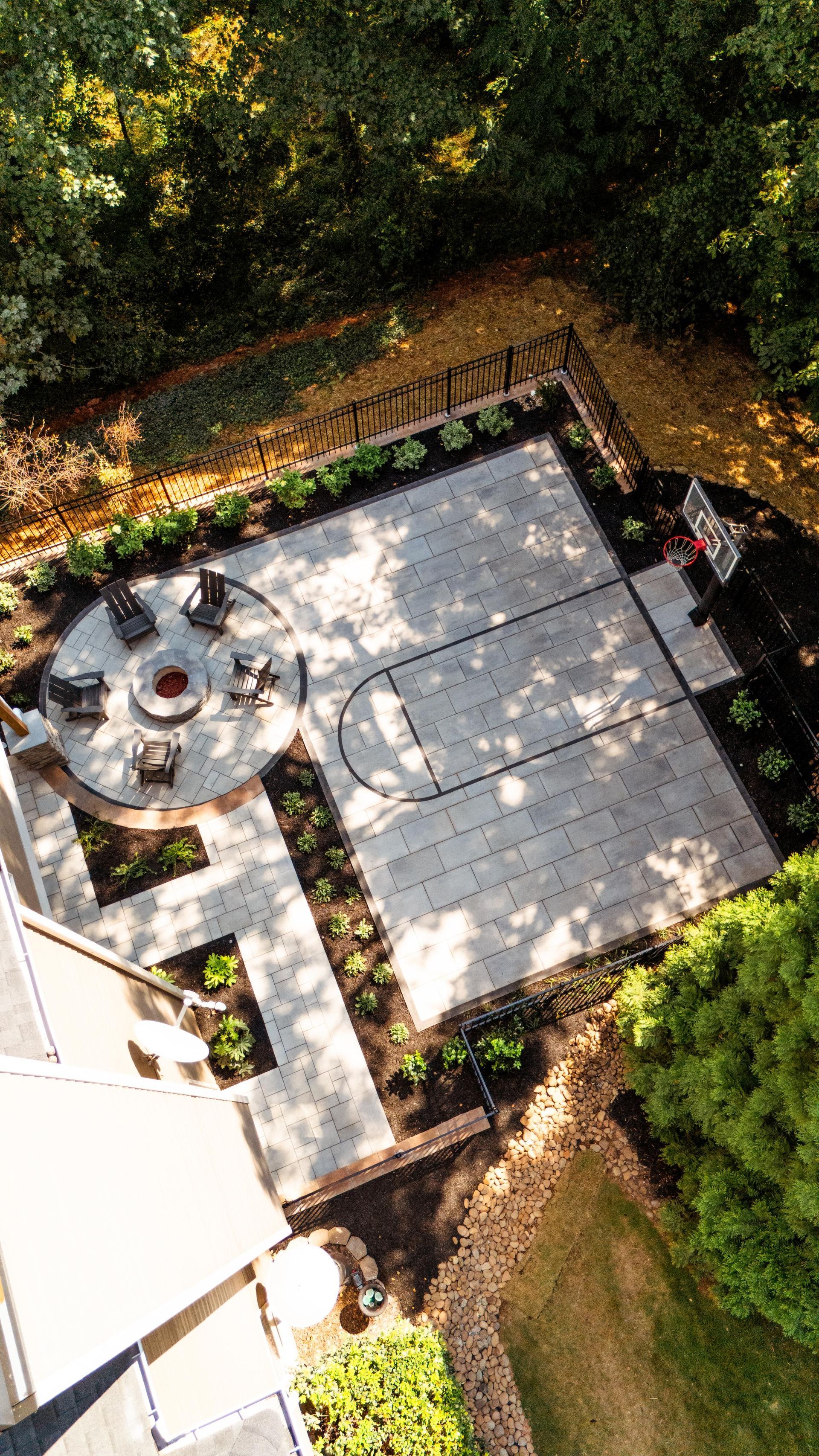 Overhead view of a concrete backyard with a basketball court, fire pit, and patio seating area.