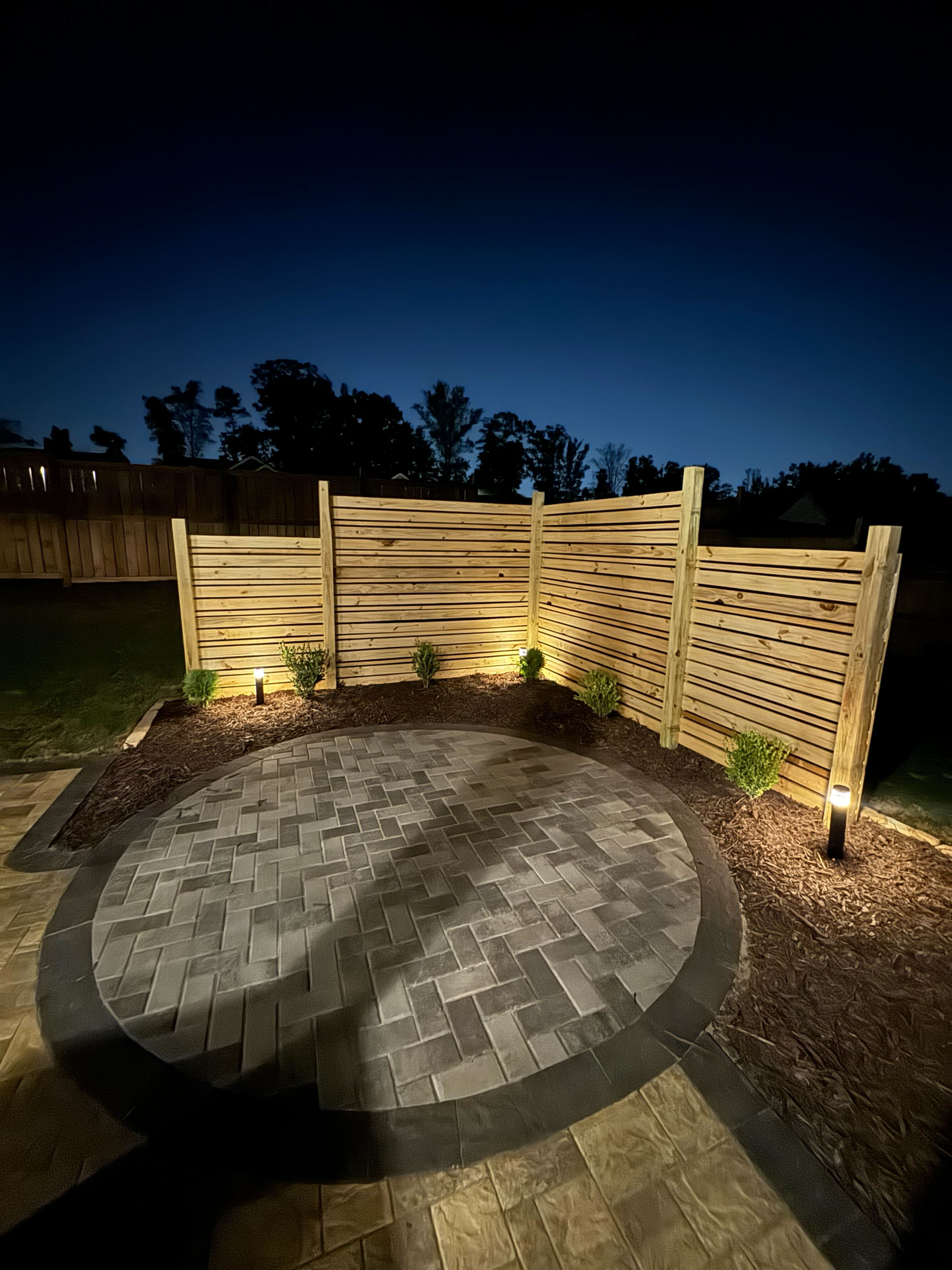 A backyard patio with a decorative wooden fence, lit by spotlights at night.