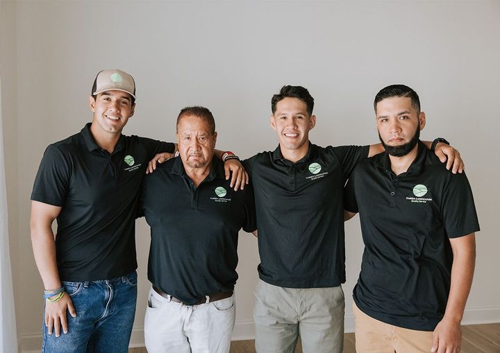 Four people in matching black polo shirts, arms around each other, smiling against a white wall.