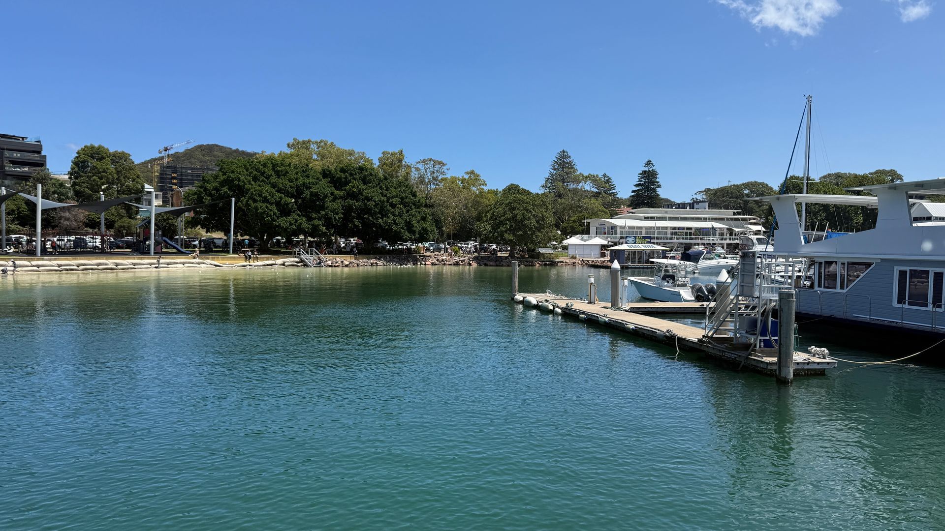Harbor scene, boats docked at pier, calm blue water, trees and buildings in background under clear sky.