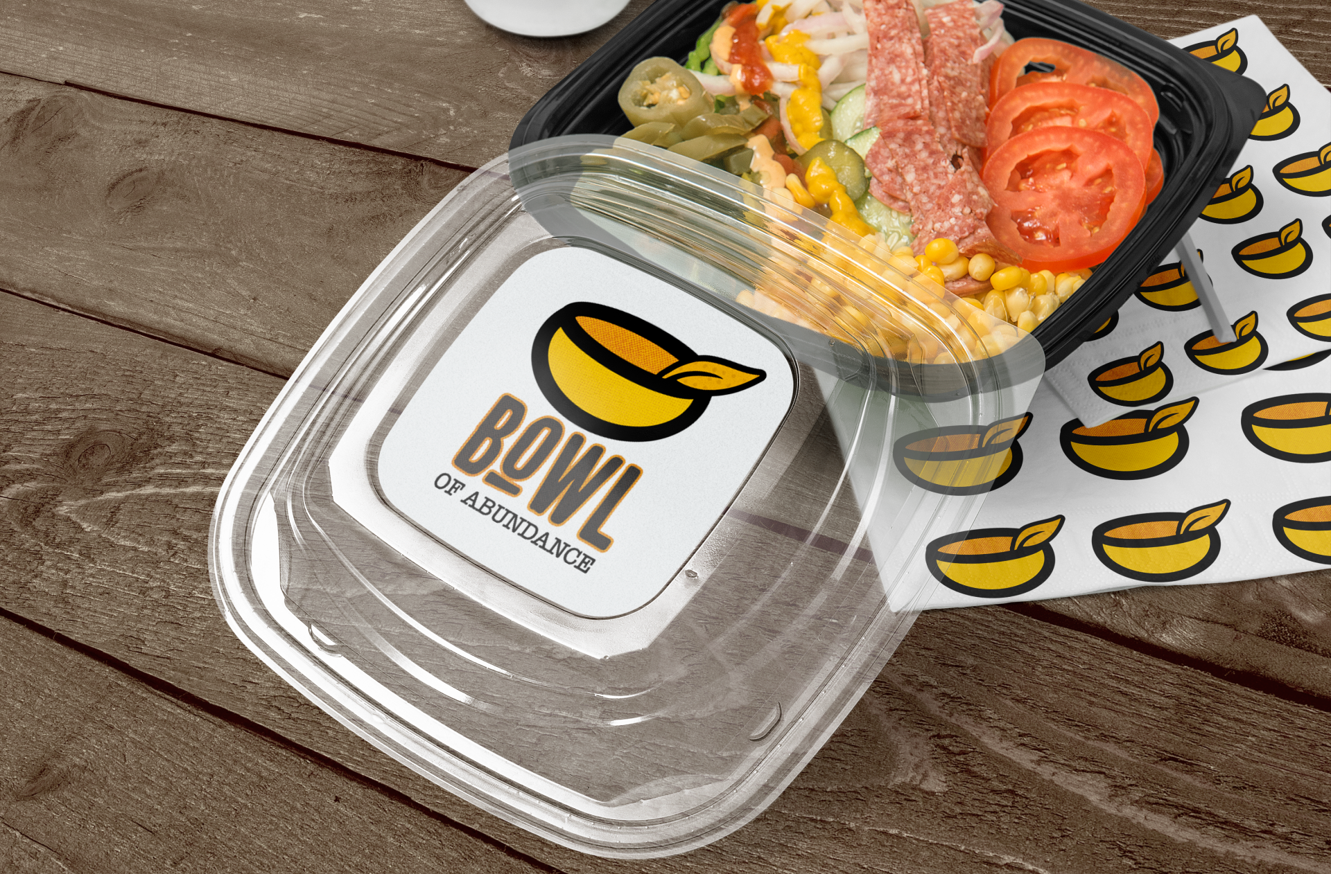 A take-out bowl with fresh ingredients sits on wood. The container features a logo with the words 