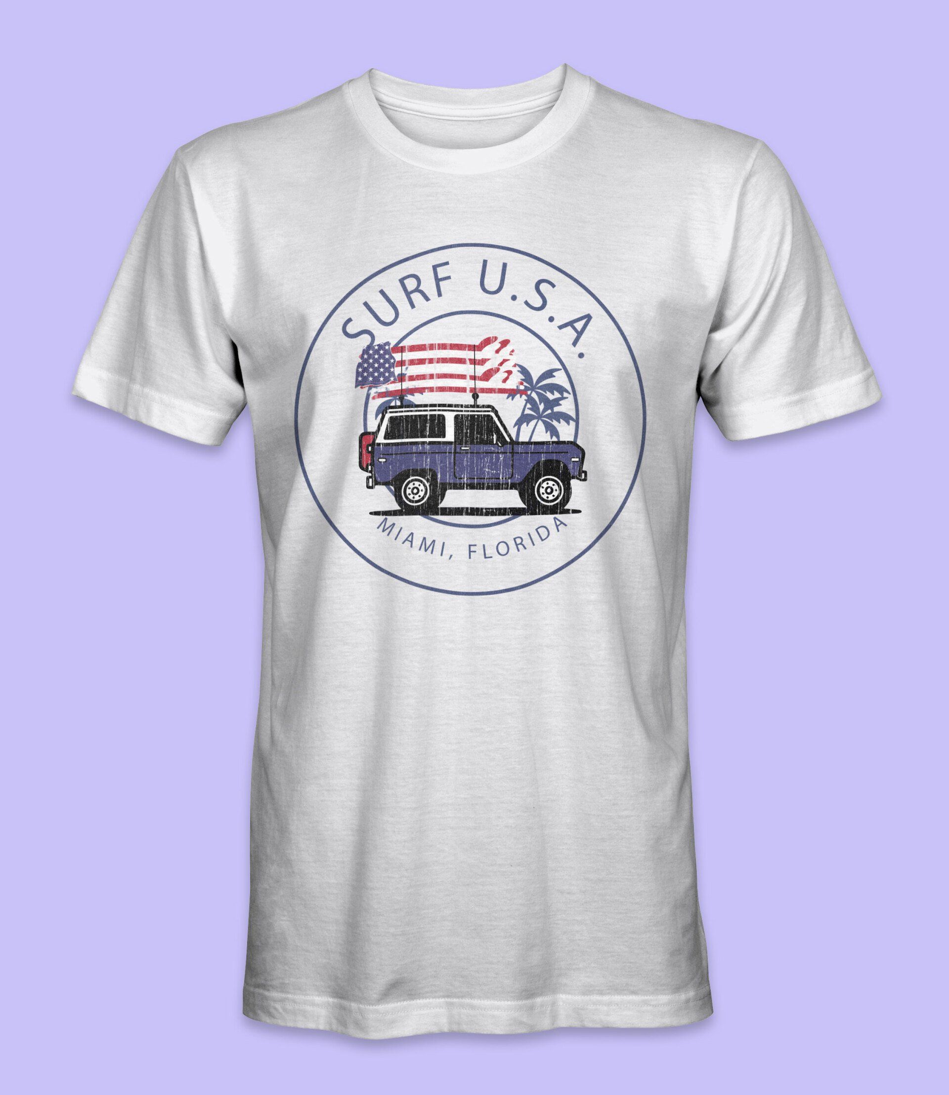 White t-shirt with a design of a blue SUV carrying an American flag and surfboard, in a circular logo reading 