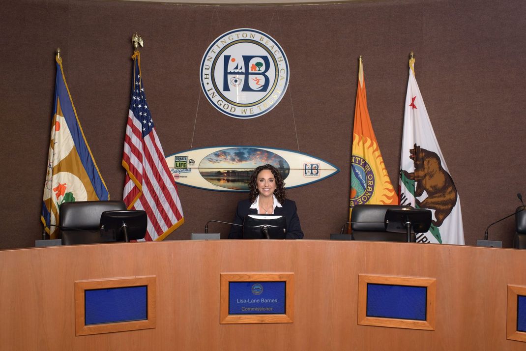 A woman sits at a desk in front of a sign that says HB.  Lisa Lane for Huntington Beach City Clerk