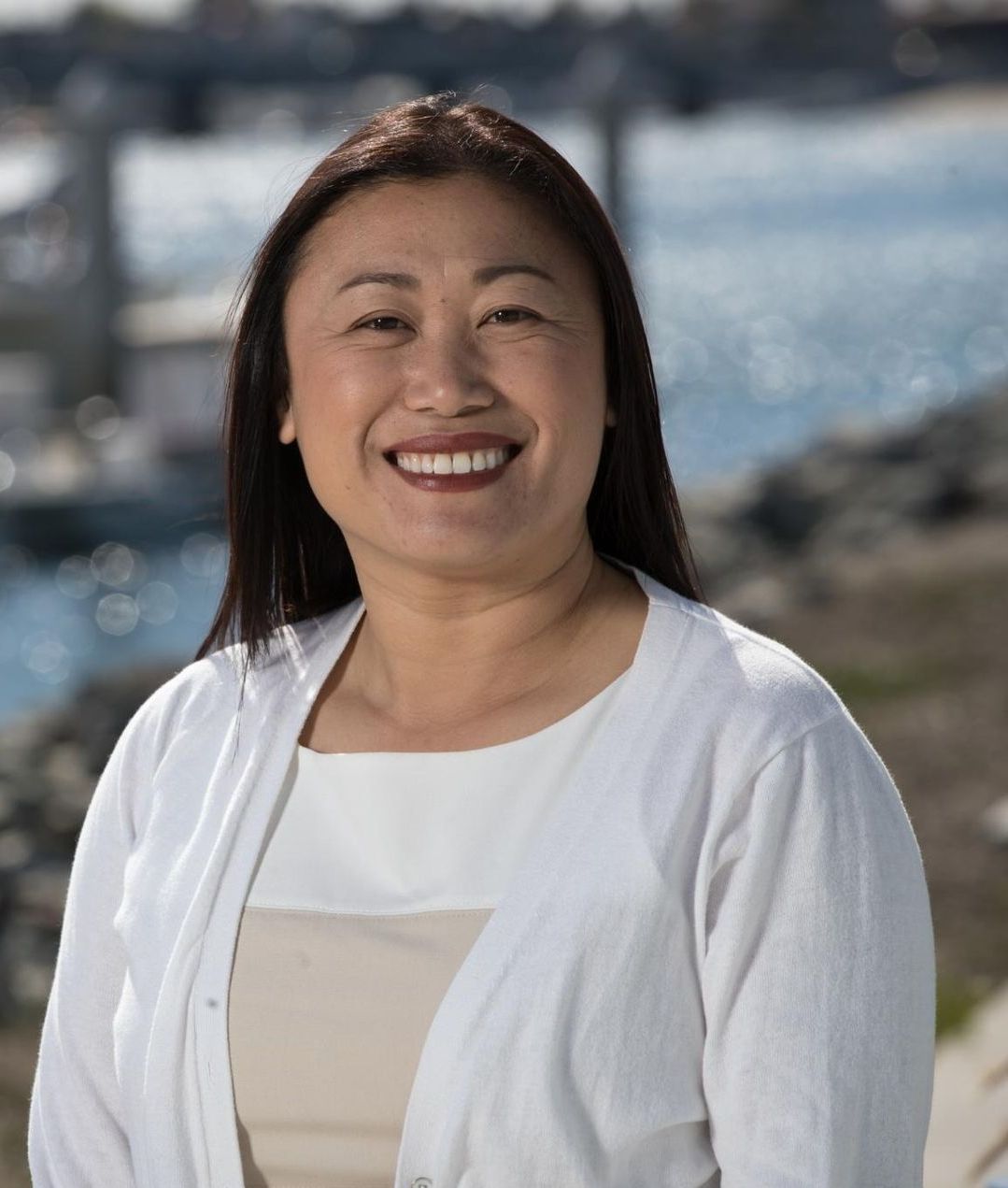 A woman in a white sweater is smiling in front of a body of water. Janet Nguyen
