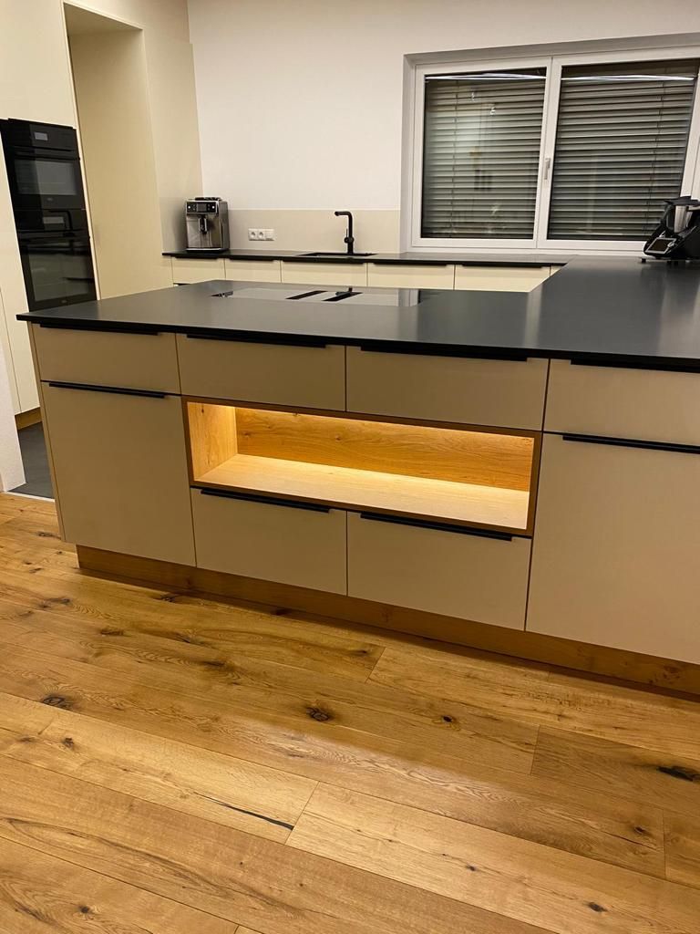 Moderne Kücheninsel mit cremefarbenen Schränken, schwarzer Arbeitsplatte und beleuchtetem, offenem Holzregal auf Holzboden.