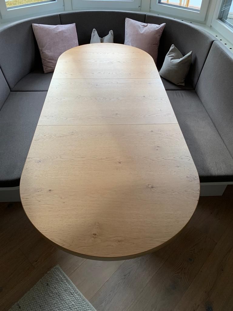 Vor einer geschwungenen, grau gepolsterten Eckbank mit vier Zierkissen steht ein ovaler Esstisch aus hellem Holz.