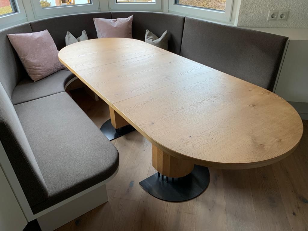 Ein ovaler Holztisch, umgeben von einer geschwungenen, braun gepolsterten Sitzbank mit hellrosa Zierkissen.
