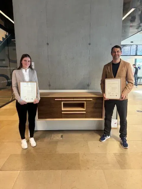 Zwei Personen mit Urkunden stehen auf beiden Seiten eines hölzernen Sideboards