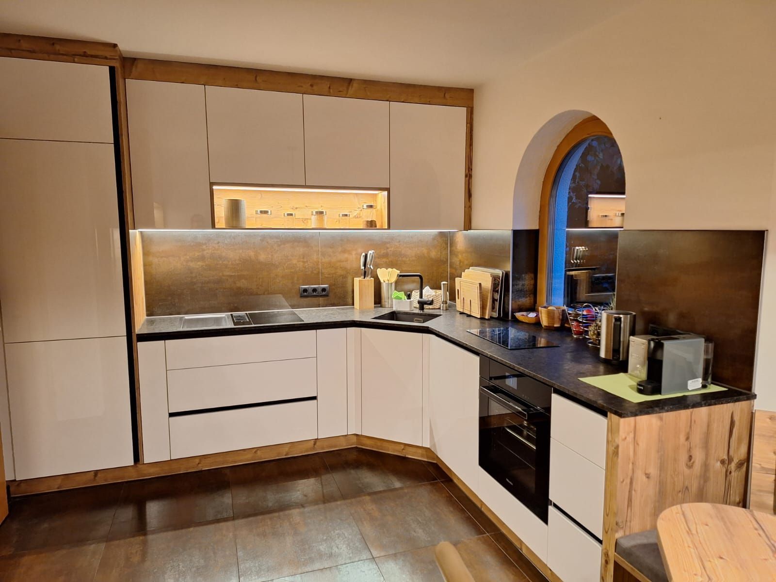 Moderne L-förmige Küche mit weißen Schränken, Holzelementen, dunklen Arbeitsplatten und einem Bogenfenster.