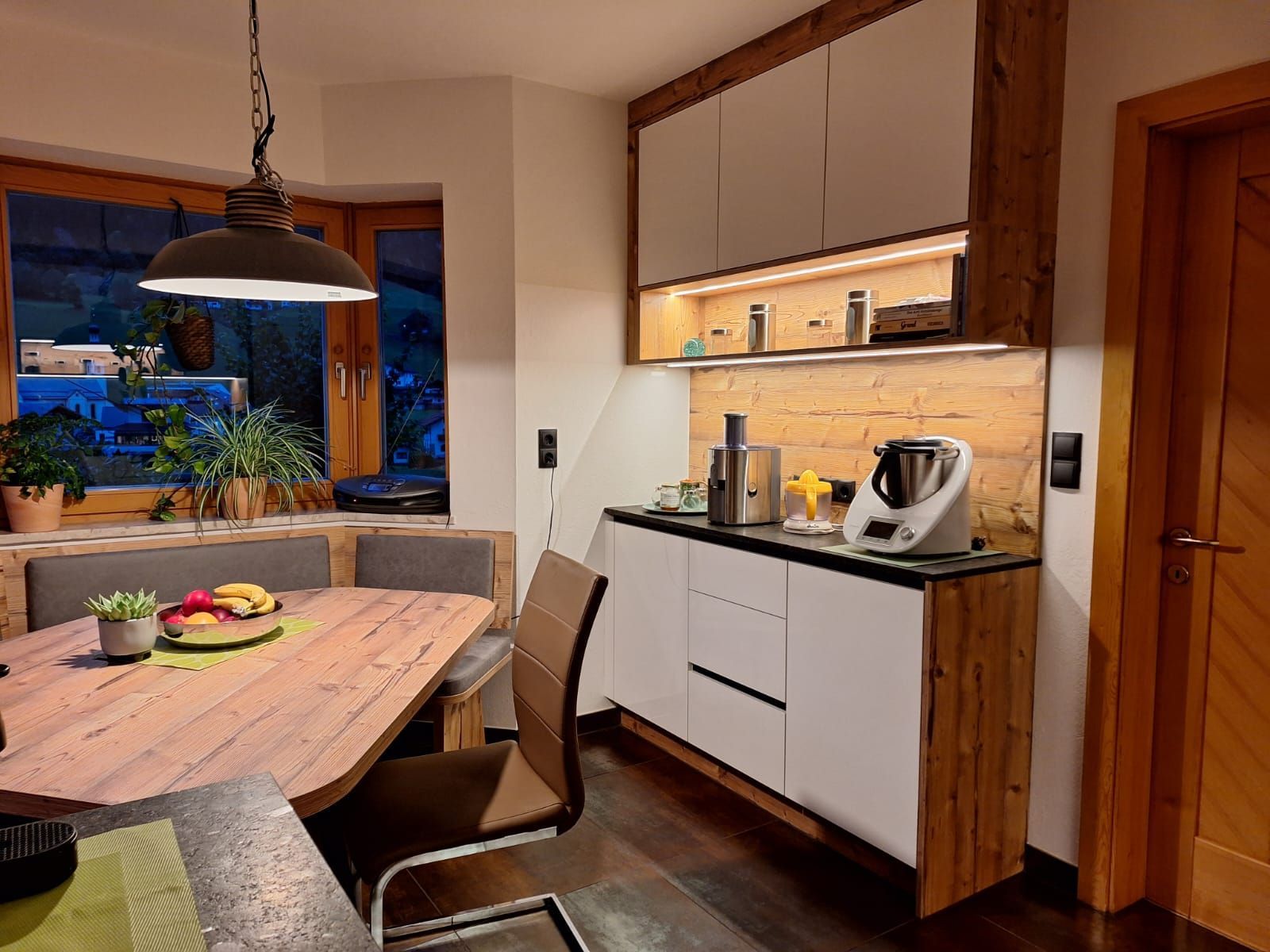 Eine gemütliche Essecke mit Holztisch, Sitzbank, Hängelampe und modernen Küchenschränken mit Elektrogeräten.