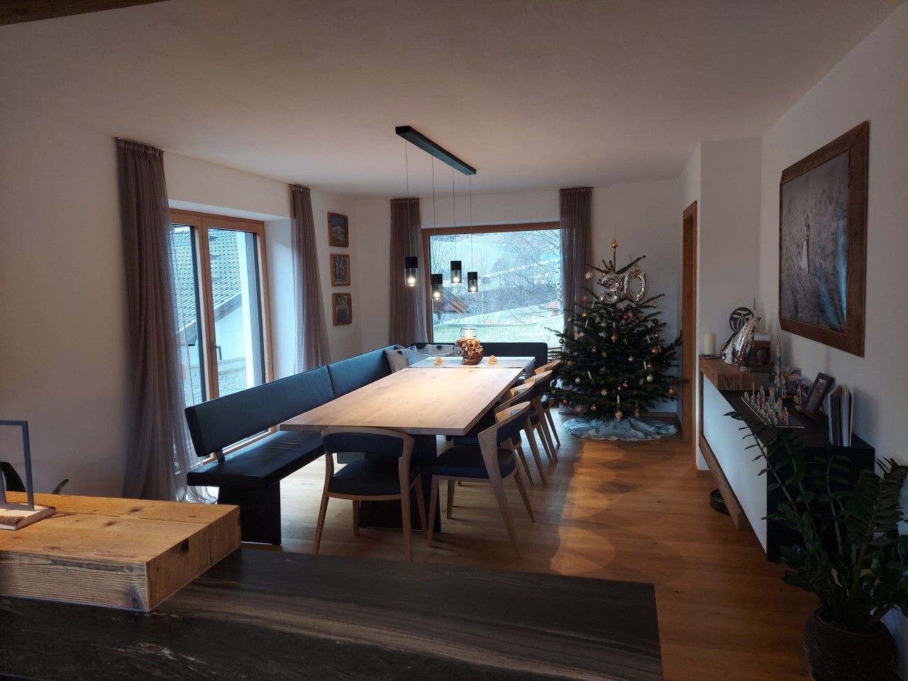 Ein Essbereich mit einem langen Holztisch, einer schwarzen Eckbank und Stühlen, in dessen Ecke ein Weihnachtsbaum steht.