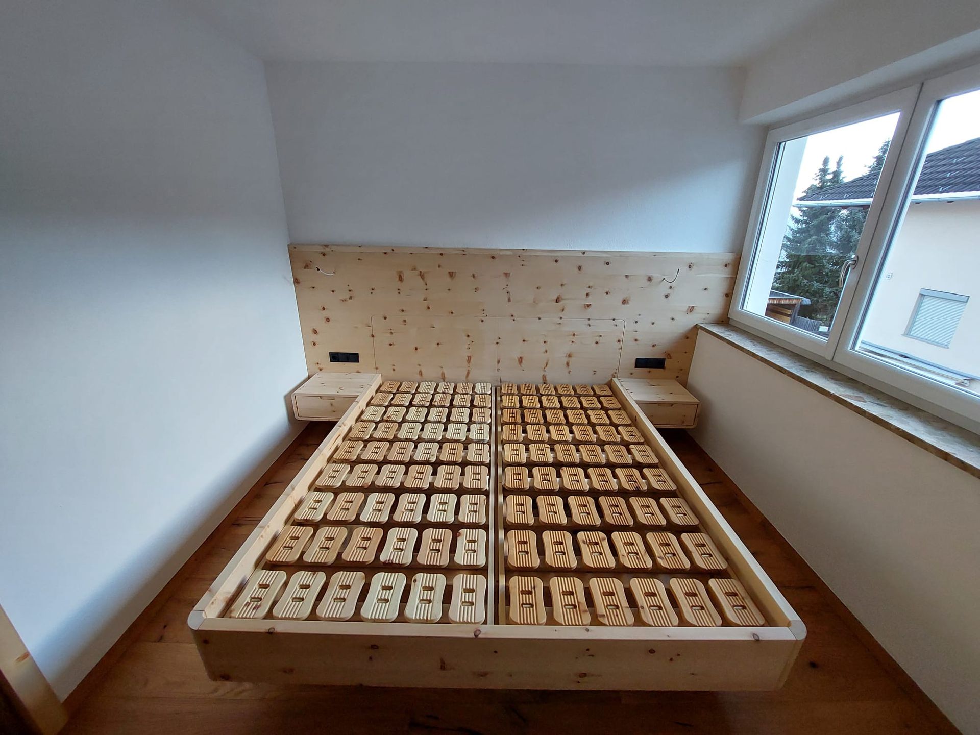 Ein helles Holzbettgestell mit integrierten Nachttischen und einem Kopfteil mit Punktmuster in einem hellen Schlafzimmer.