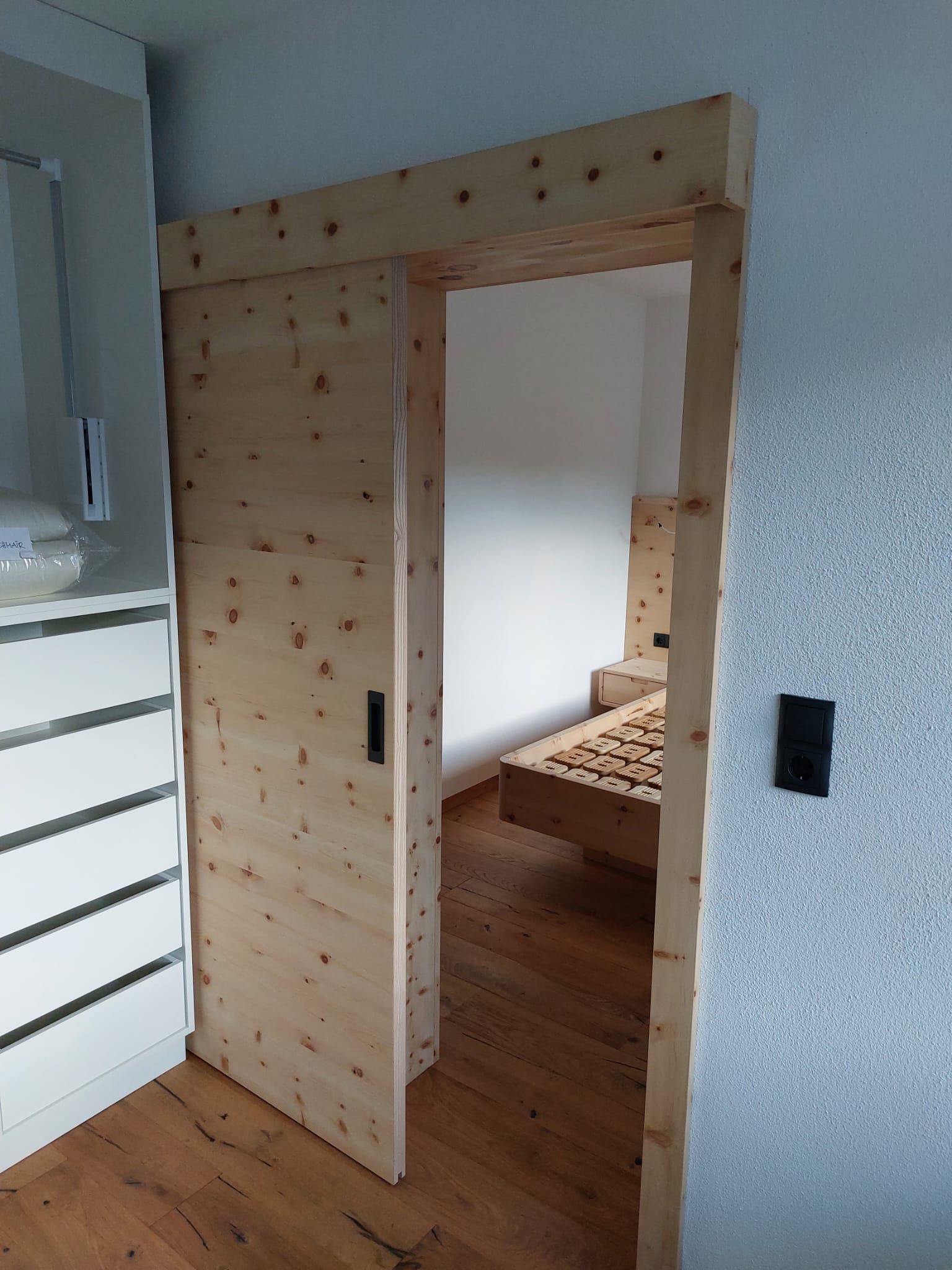 Eine helle Schiebetür aus astiger Kiefer öffnet sich zu einem Schlafzimmer mit einem Bettgestell und Holzfußboden.