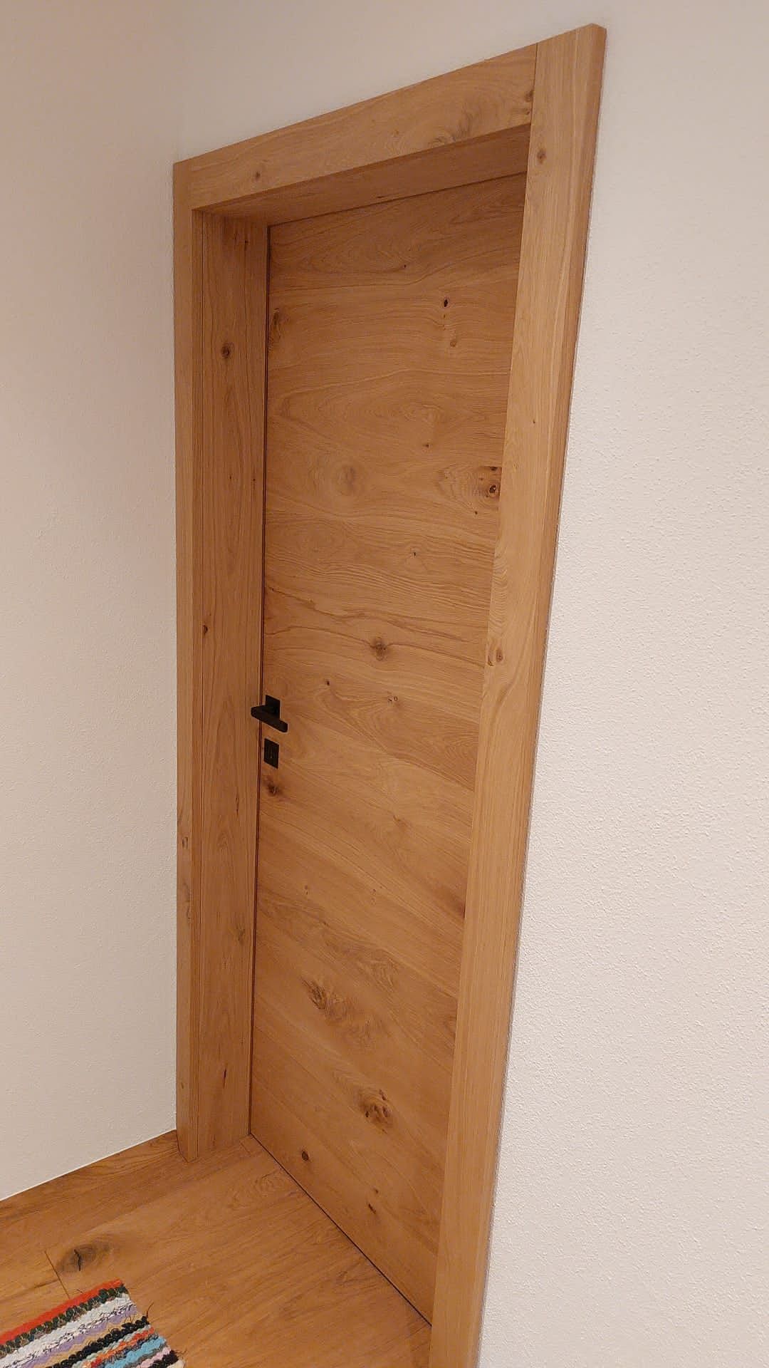 Eine helle Holztür mit schwarzem Griff, eingefasst in einen passenden Holzrahmen vor schlichten Wänden.