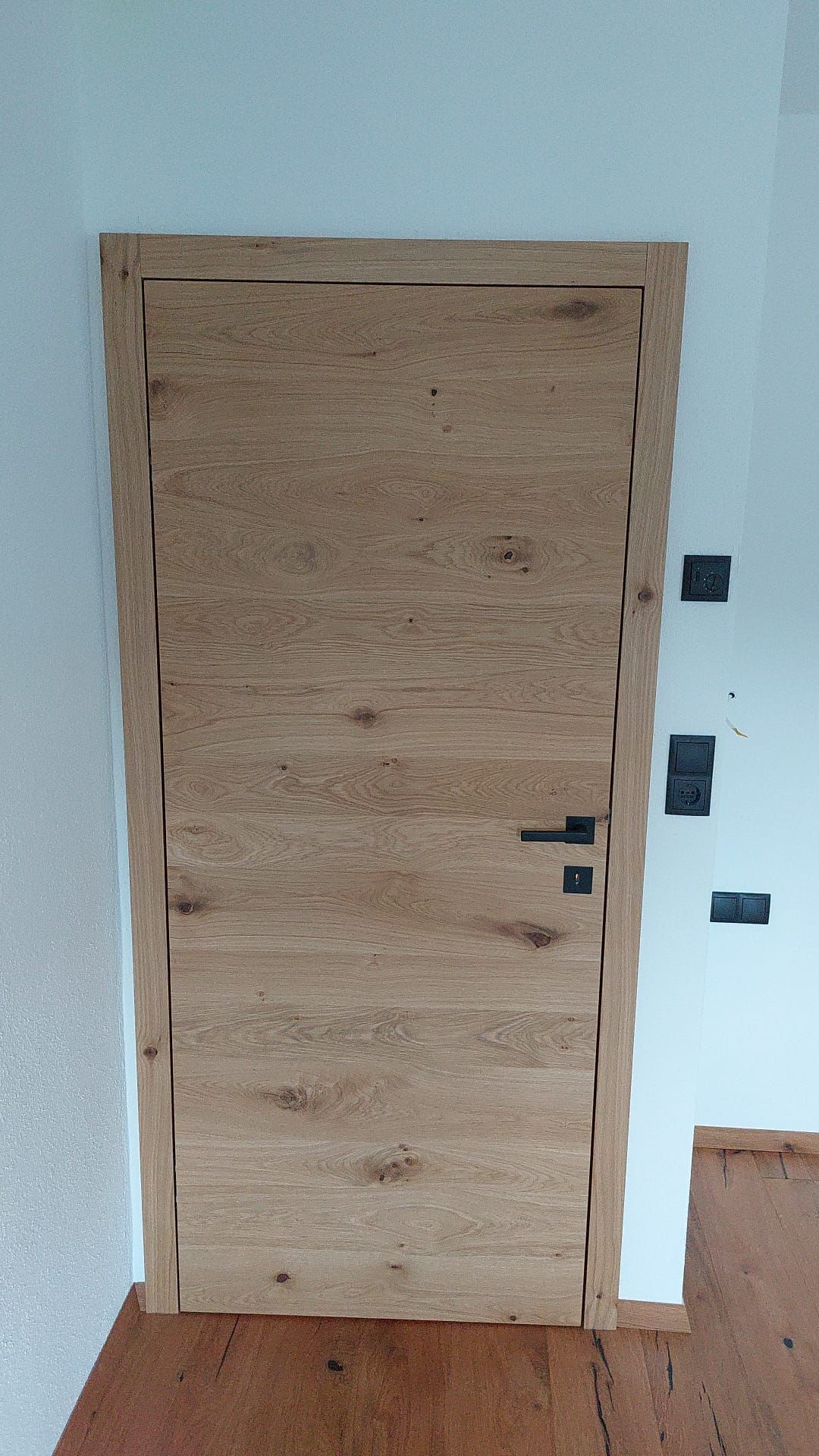 Eine rustikale Holztür mit horizontaler Maserung, eingefasst in eine weiße Wand mit dunklen Beschlägen und einem Holzboden.