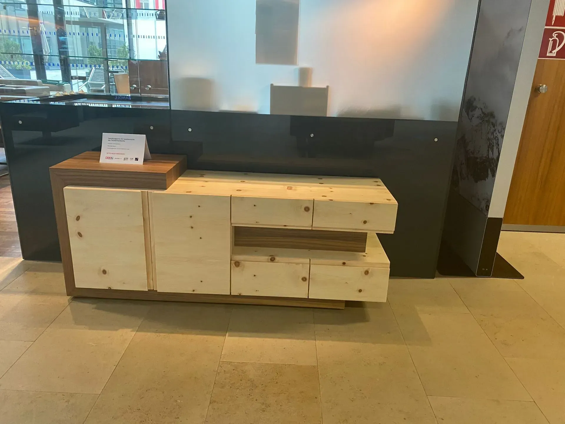 Ein helles Holz-Sideboard mit zwei Türen und vier Schubladen, das in einer Lobby an einer dunkelgrauen Theke steht.