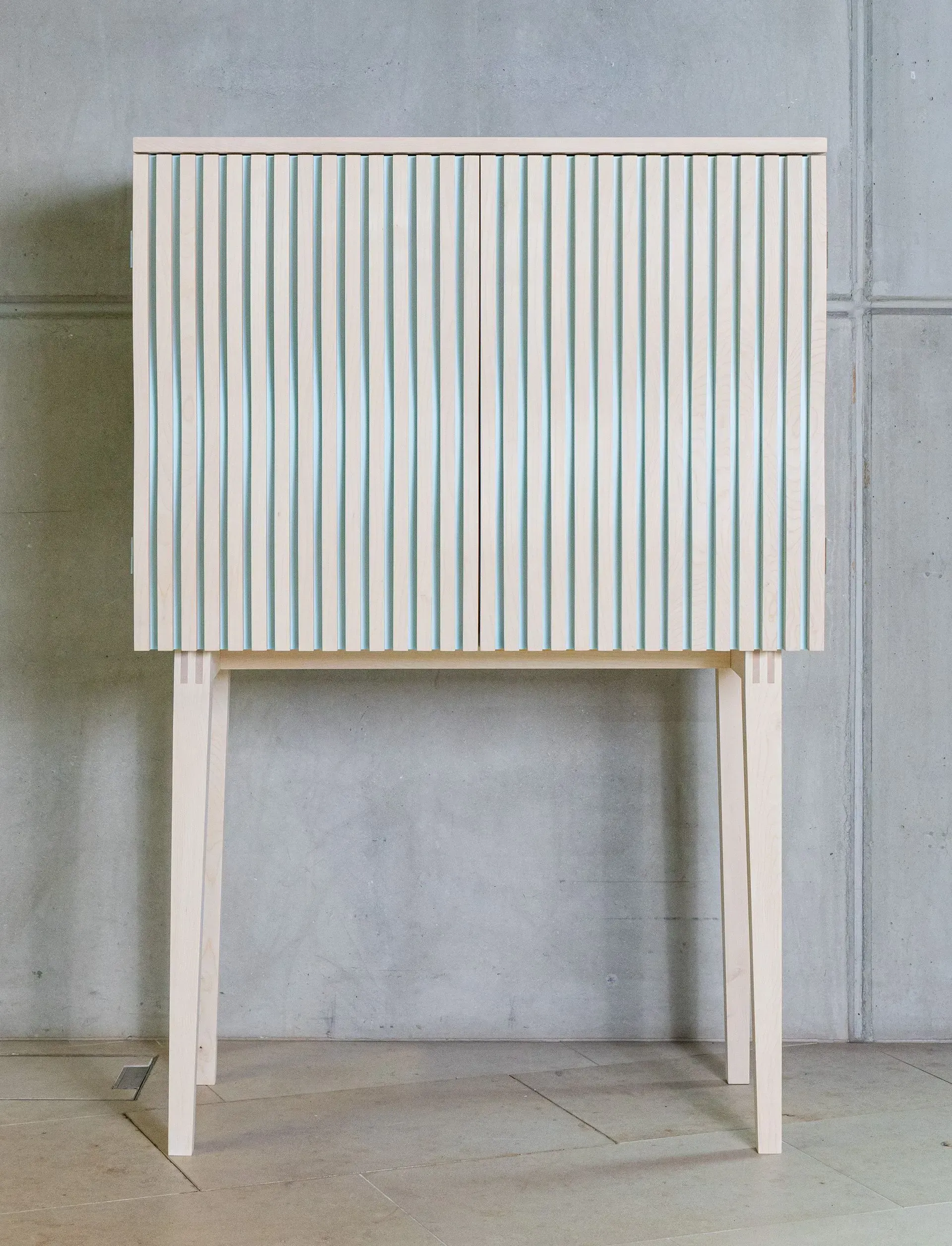 Ein heller Holzschrank auf dünnen Beinen mit vertikal geriffelten Türen vor einem grauen Betonhintergrund.