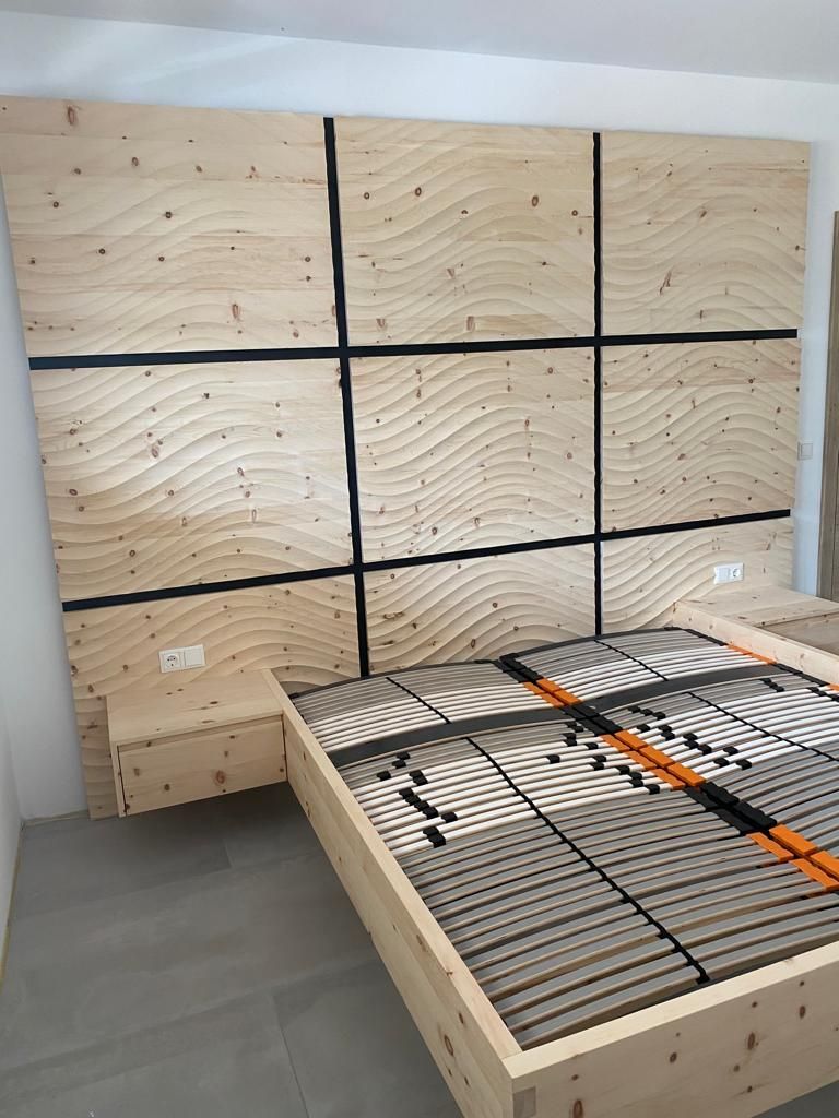 Ein Bett mit Lattenrost, das an einem großen, dekorativen Wandkopfteil mit hellen Holzpaneelen und Wellenmuster steht.