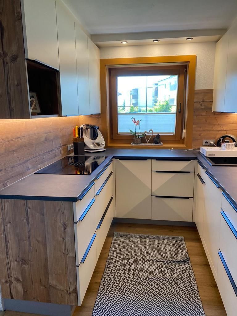 Moderne U-förmige Küche mit weißen Schränken, Akzenten in Holzoptik, dunklen Arbeitsplatten und einem Fenster über der Spüle.