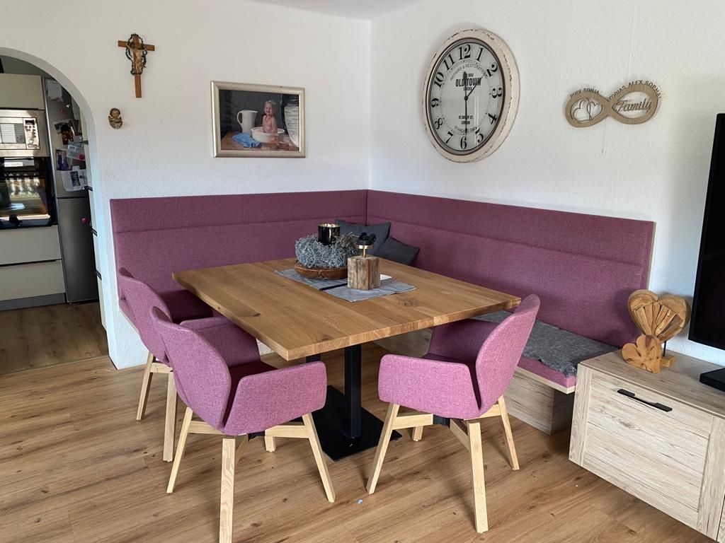 Ein Essbereich mit einem Holztisch, einer lila Sitzbank und zwei passenden Sesseln auf einem hellen Holzboden.