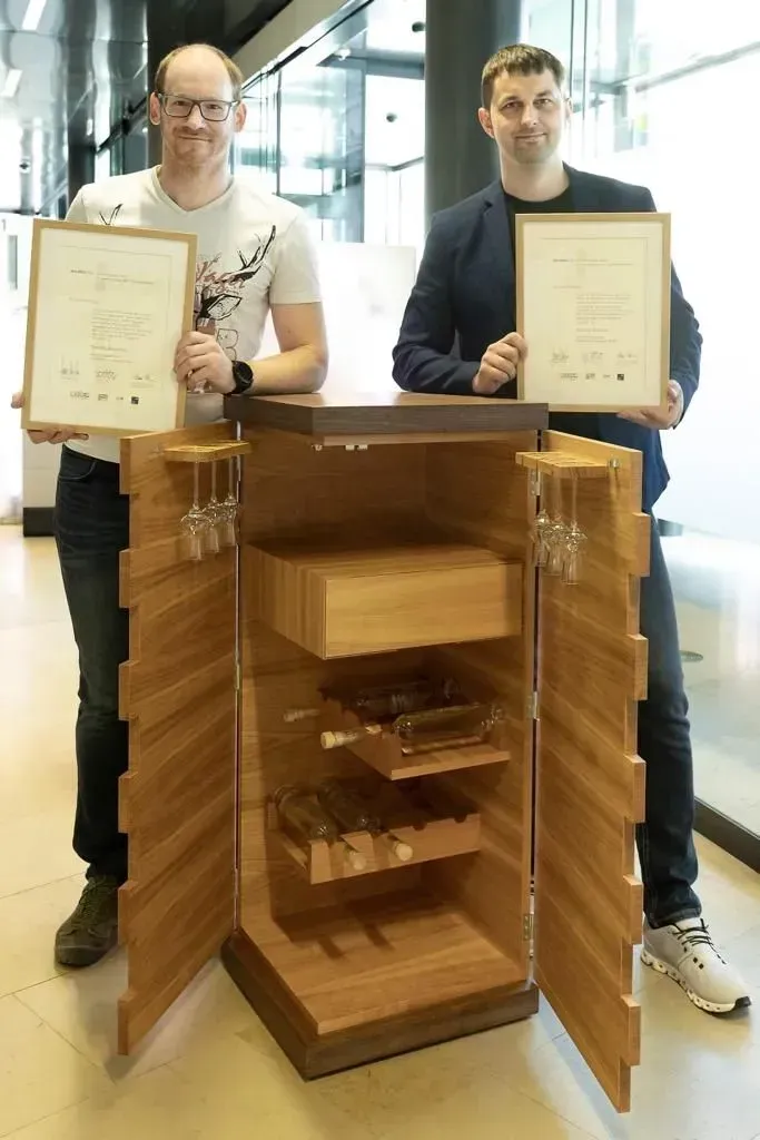 In einem hell erleuchteten Büro stehen zwei Personen hinter einem Holzschrank, in dem gerahmte Urkunden aufbewahrt werden.