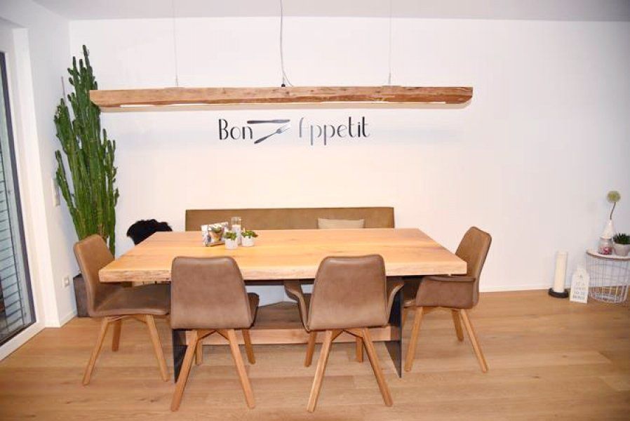 Ein Esstisch aus Holz mit passender Bank und vier braunen Stühlen unter einer rustikalen Holzbalkenleuchte.