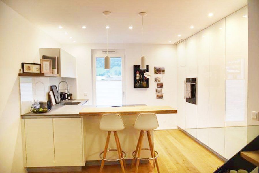 Eine moderne weiße Küche mit hölzerner Frühstückstheke, zwei Barhockern, Deckenbeleuchtung und hellem Holzboden.