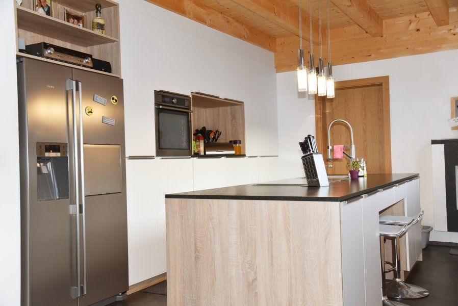 Eine moderne Küche mit einer hellen Holzinsel, einem hohen Edelstahlkühlschrank und Pendelleuchten unter Holzbalken.