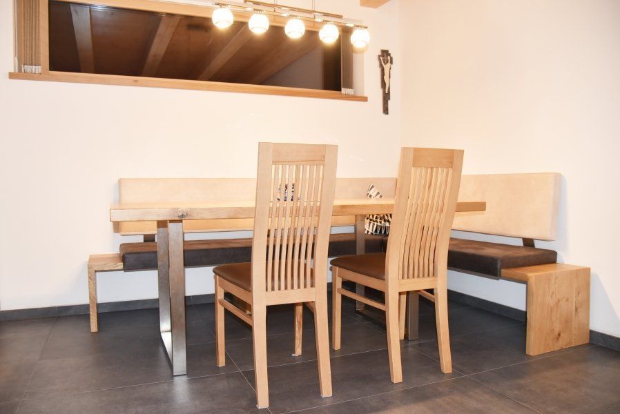 Ein hölzerner Esstisch mit zwei Stühlen und einer Eckbank mit dunklen Kissen steht vor einer weißen Wand mit einem Fenster.