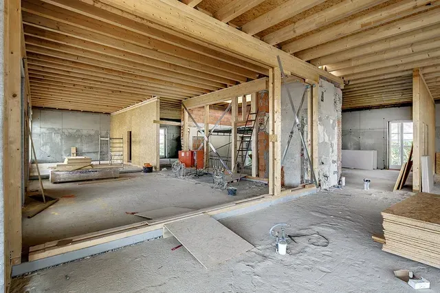 Innenansicht eines im Bau befindlichen Gebäudes mit freiliegenden Holzbalken an der Decke und unfertigen Betonwänden.