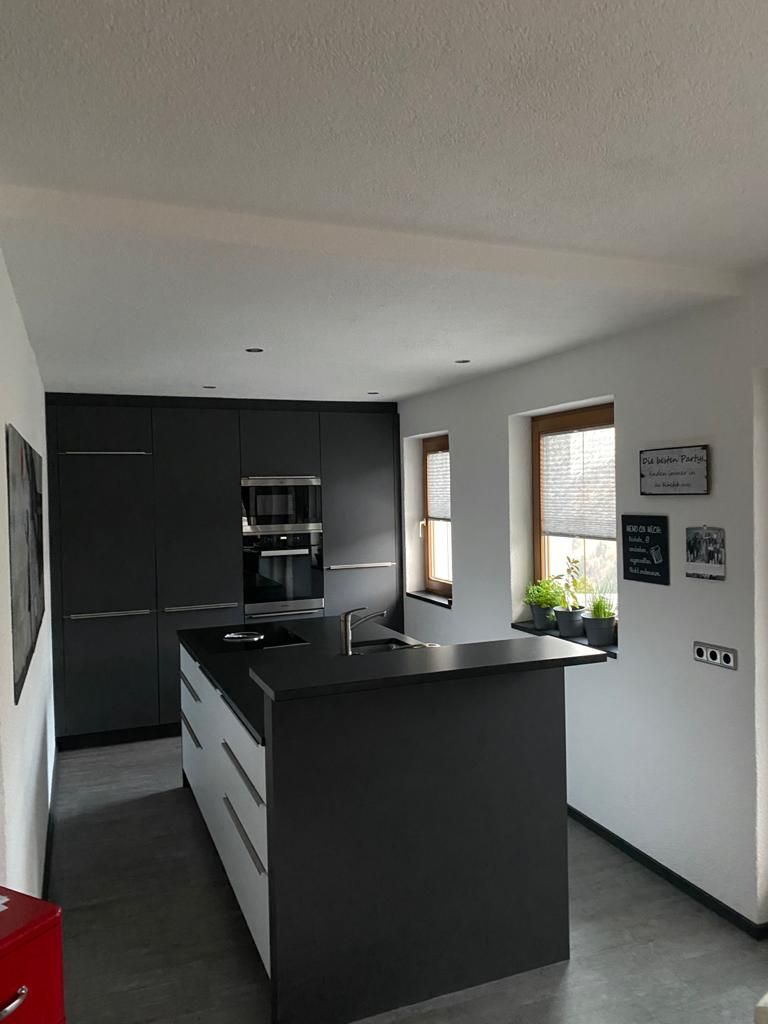 Eine moderne Küche mit schwarzer Kochinsel, dunklen Schränken, weißen Wänden und zwei Fenstern mit kleinen Topfpflanzen.