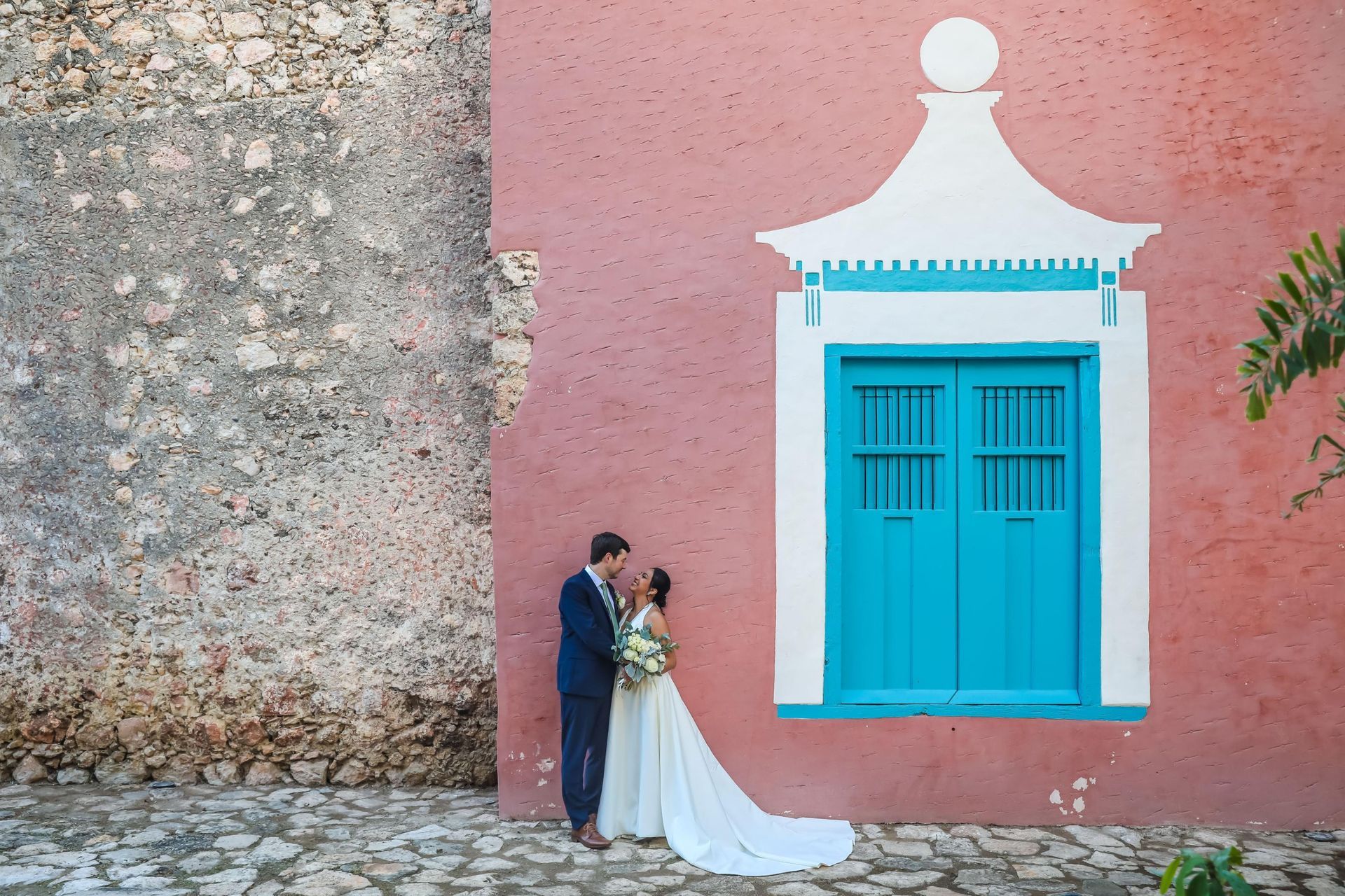 A bride and groom are kissing in front of a pink building with a blue door.